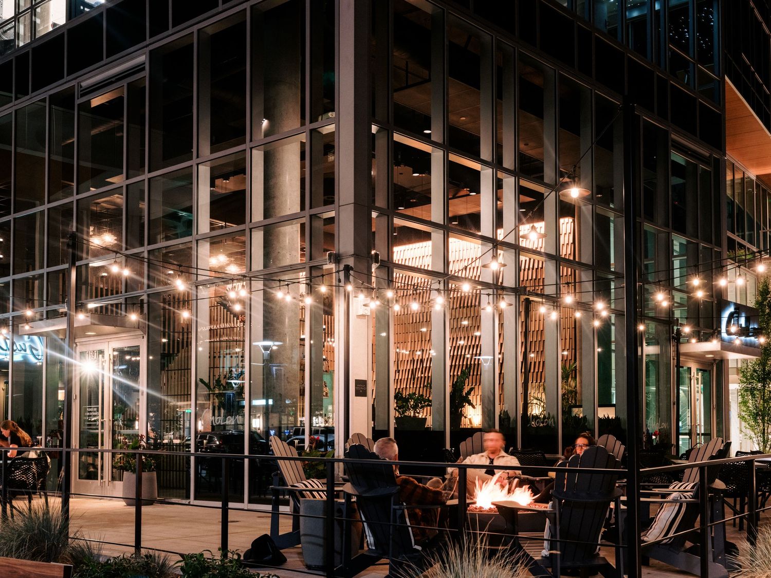 Modern restaurant exterior with glass walls and outdoor seating illuminated at night.