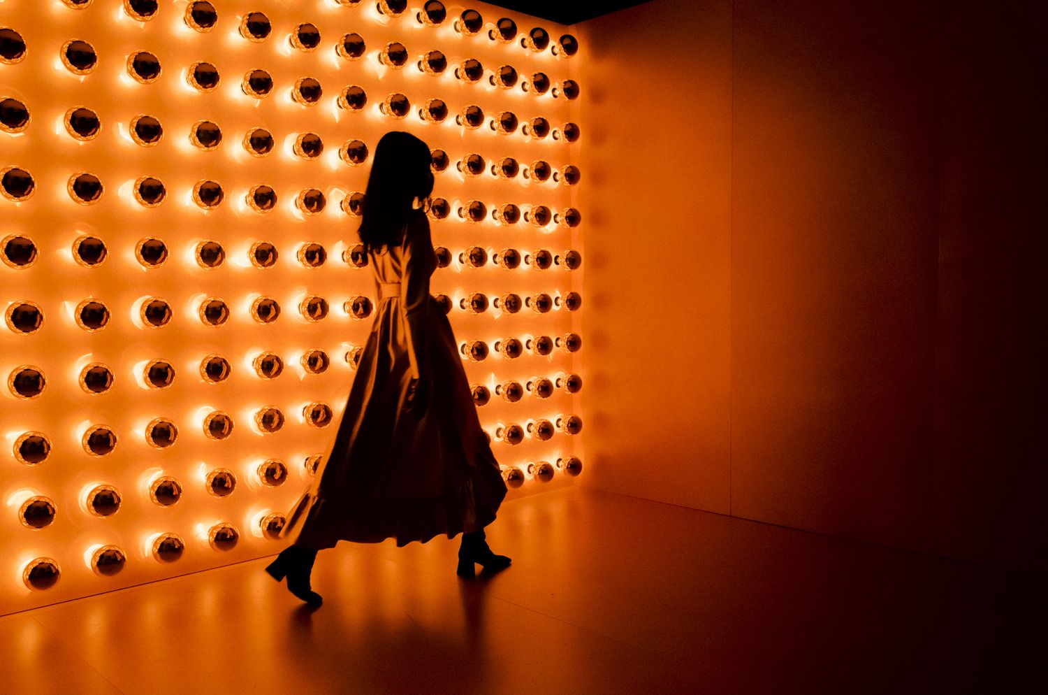 Silhouette of a woman in a flowing dress walking past a glowing orange wall of circular lights.