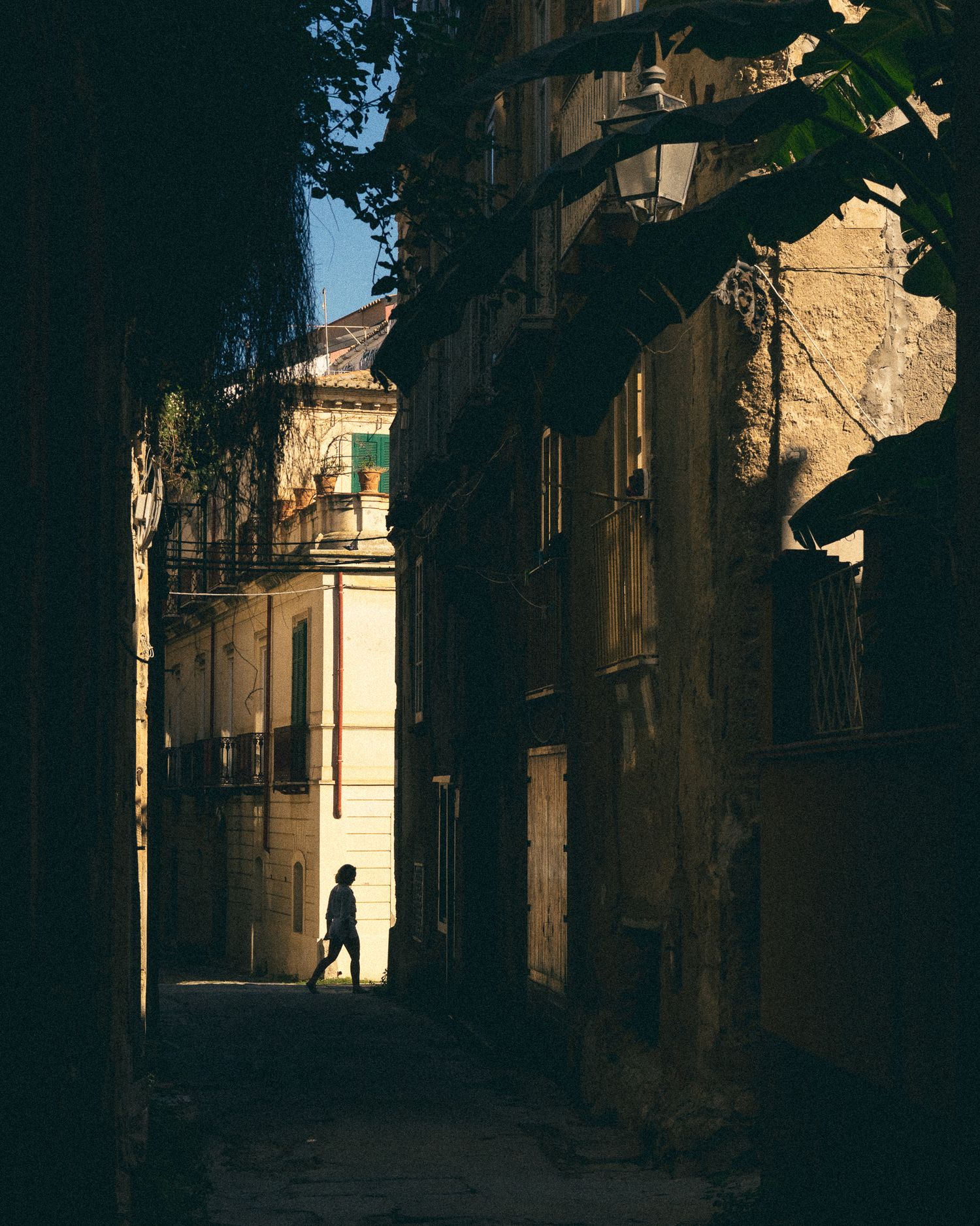 A silhouetted figure walks through a narrow, sunlit alley between old stone buildings creating dramatic shadows.