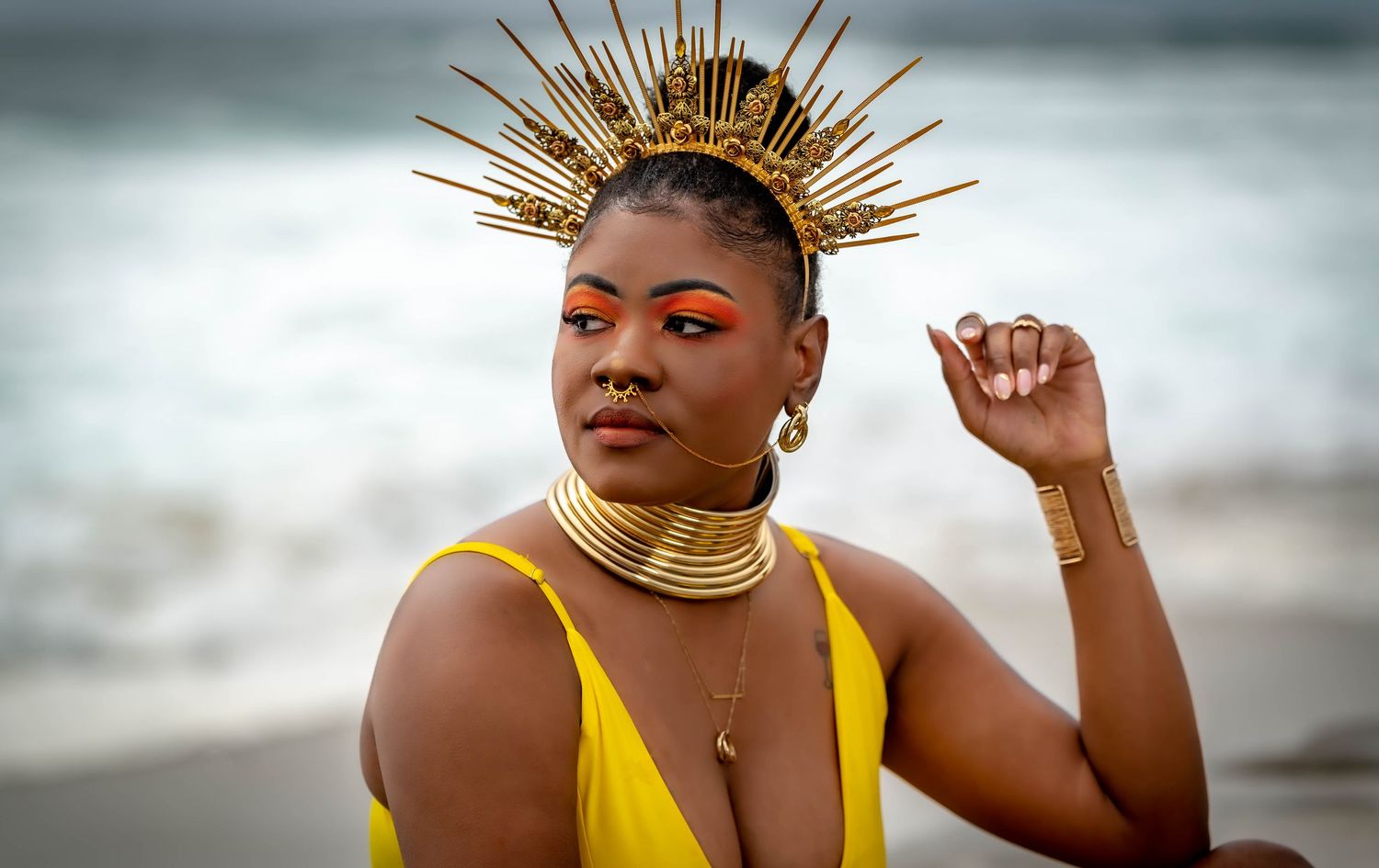 Person in yellow dress and gold accessories poses against dramatic ocean backdrop.