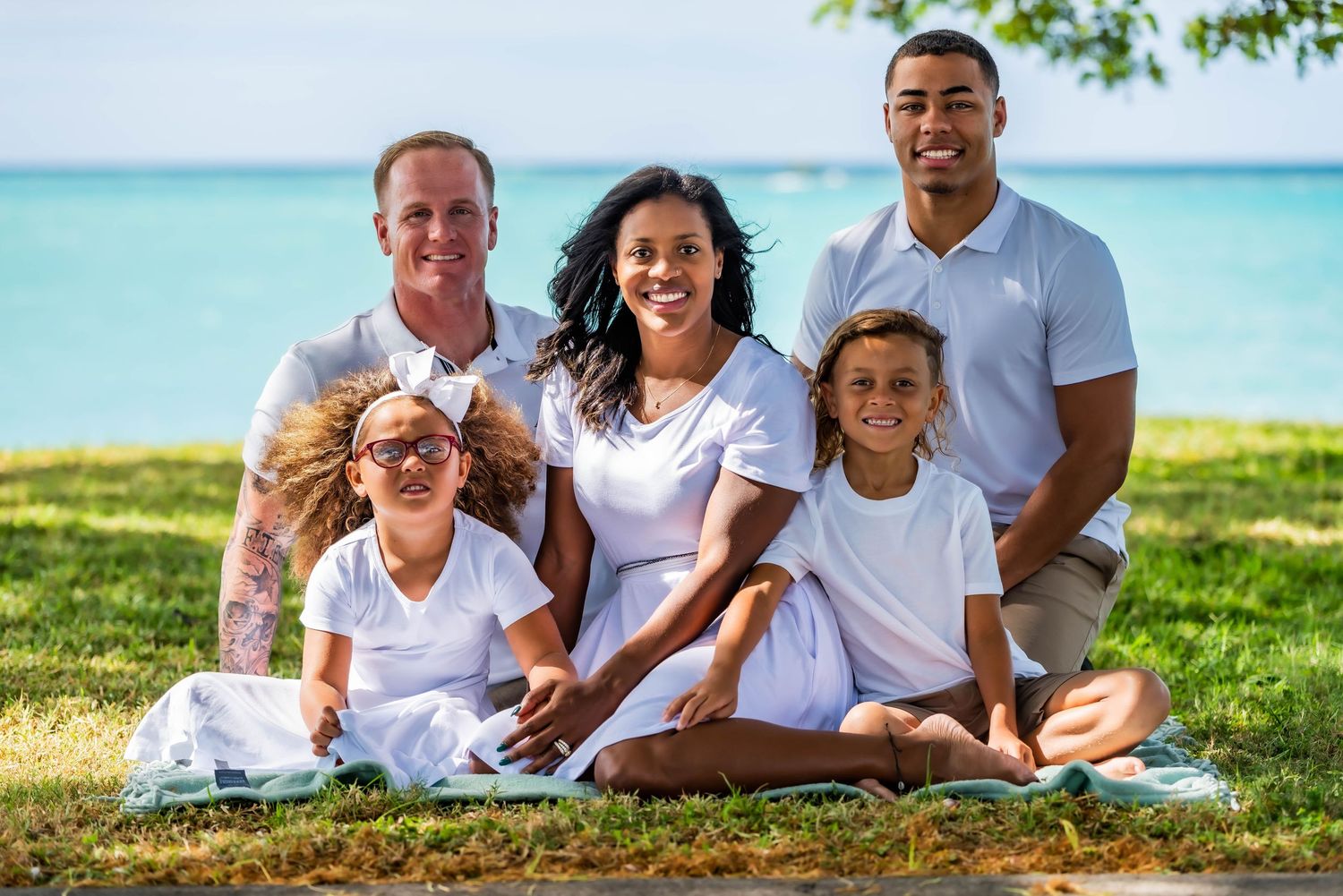 Family relaxing together on tropical beach with turquoise water in background.