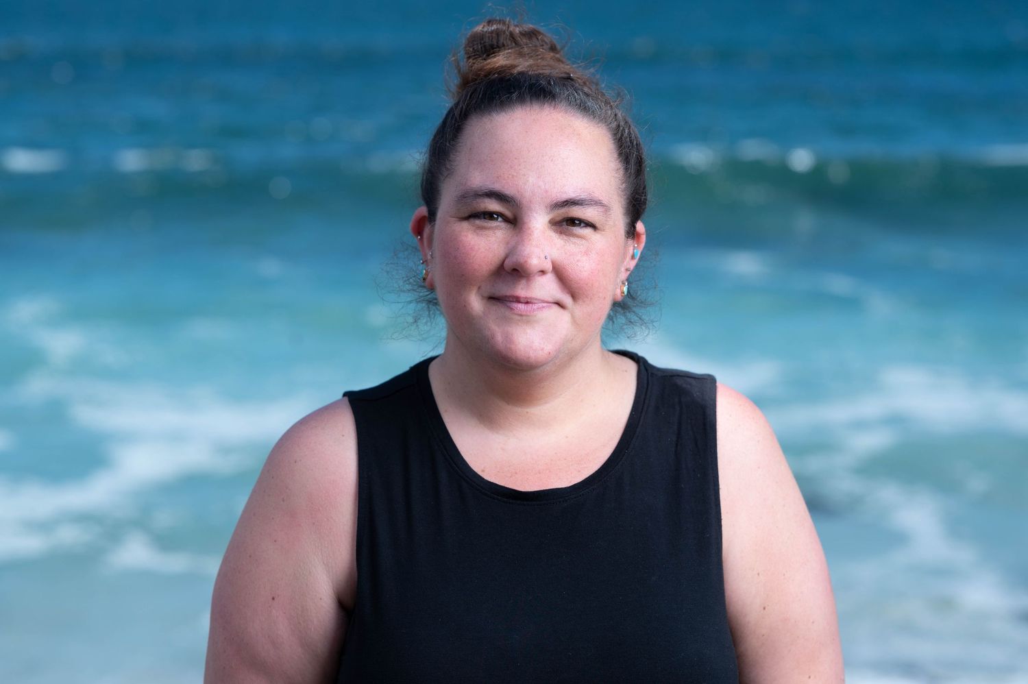 Person in black top smiling against blue ocean backdrop on sunny day.