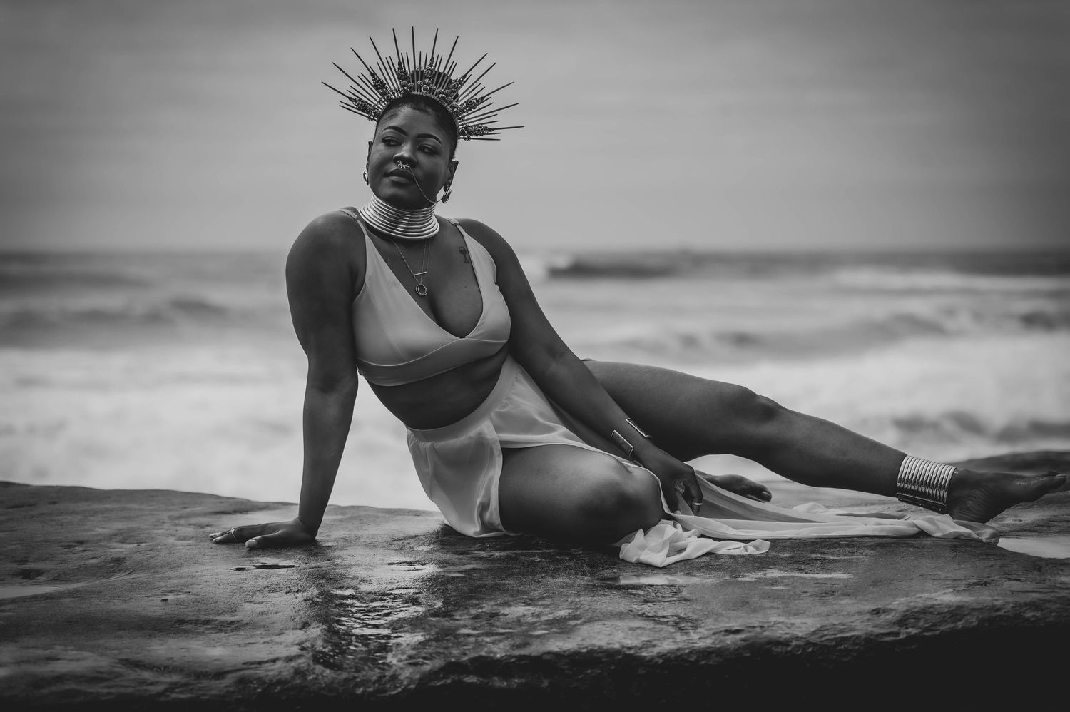 Person with spiked headpiece reclines on rocky beach in artistic black and white photo.