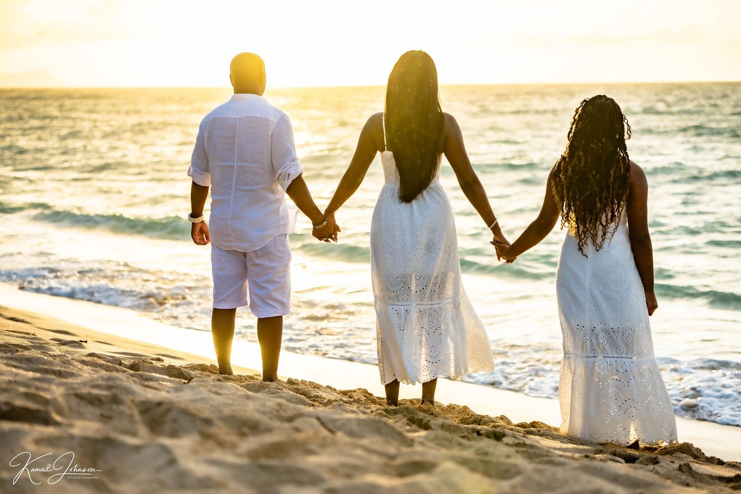 Three people hold hands while walking along beach at sunset in white attire.