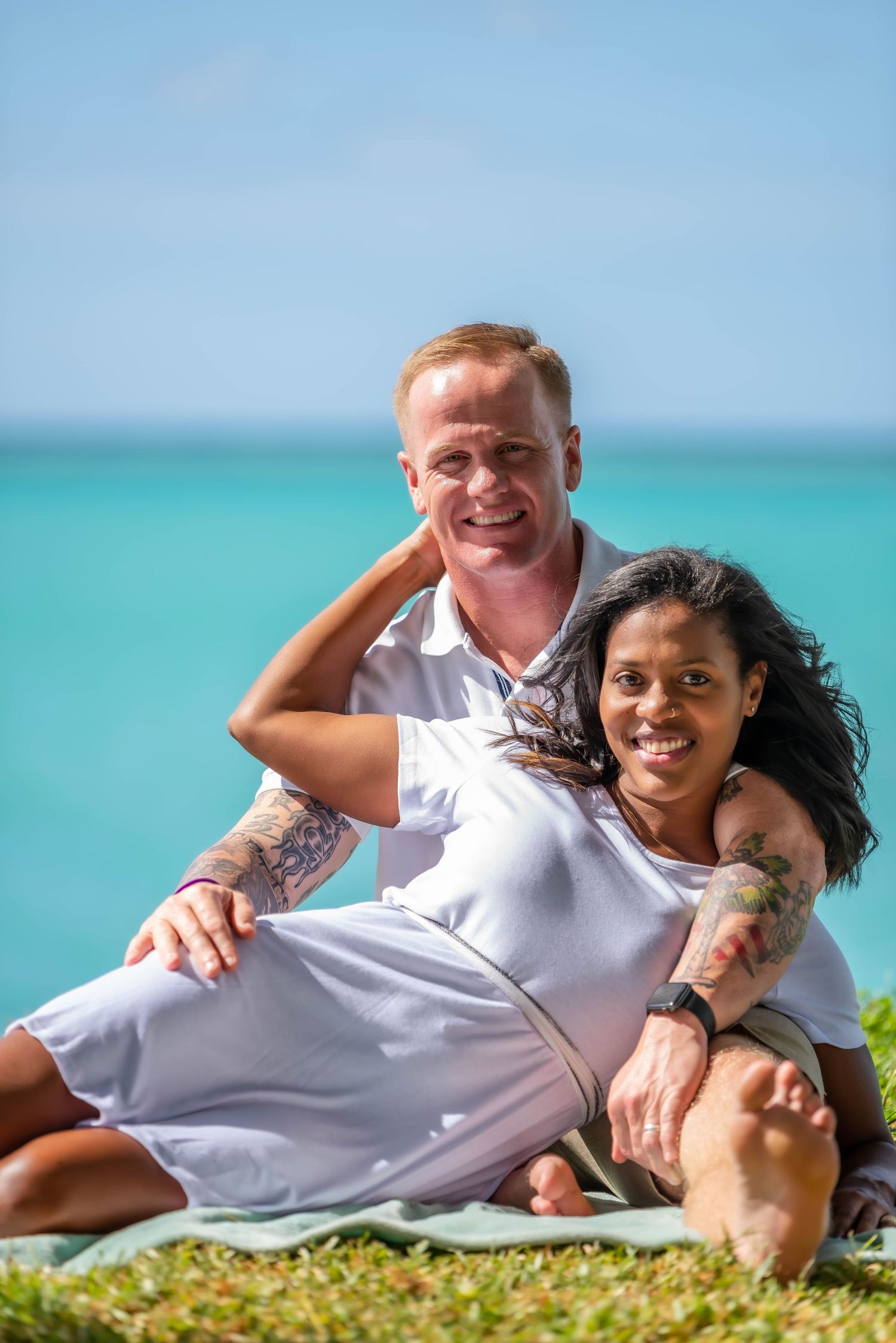 Couple in white shirts lounging on grass by turquoise ocean.