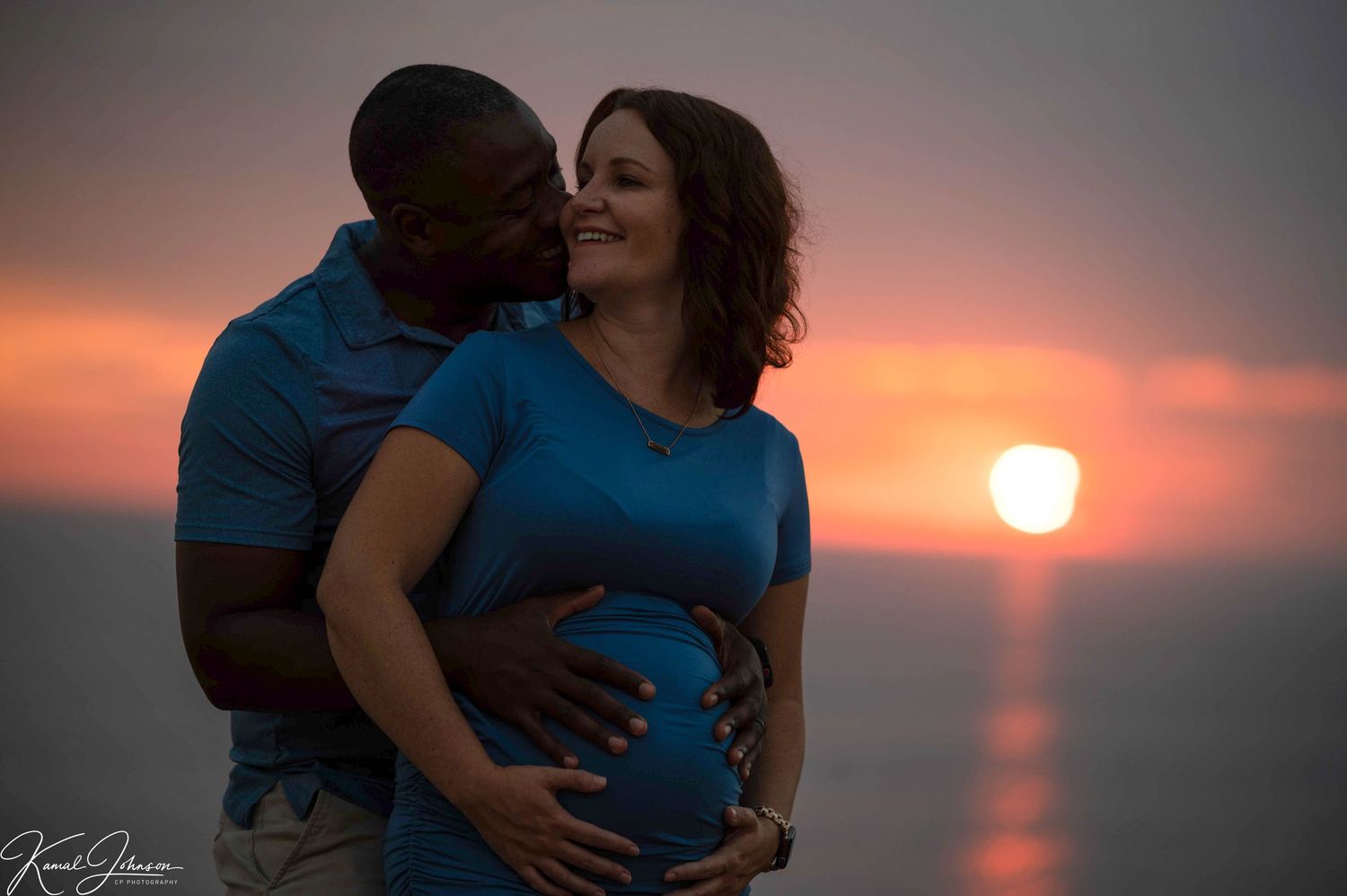 Silhouetted couple embraces during sunset maternity photoshoot on beach with glowing orange sun on horizon.