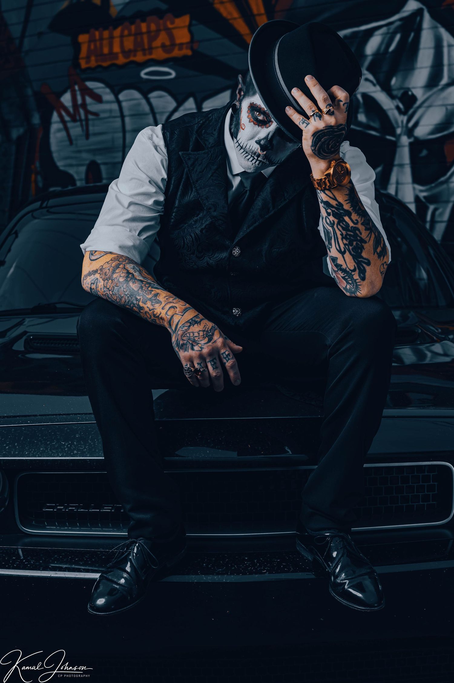 Person in dark clothing with tattoos wearing skull mask against graffiti wall.