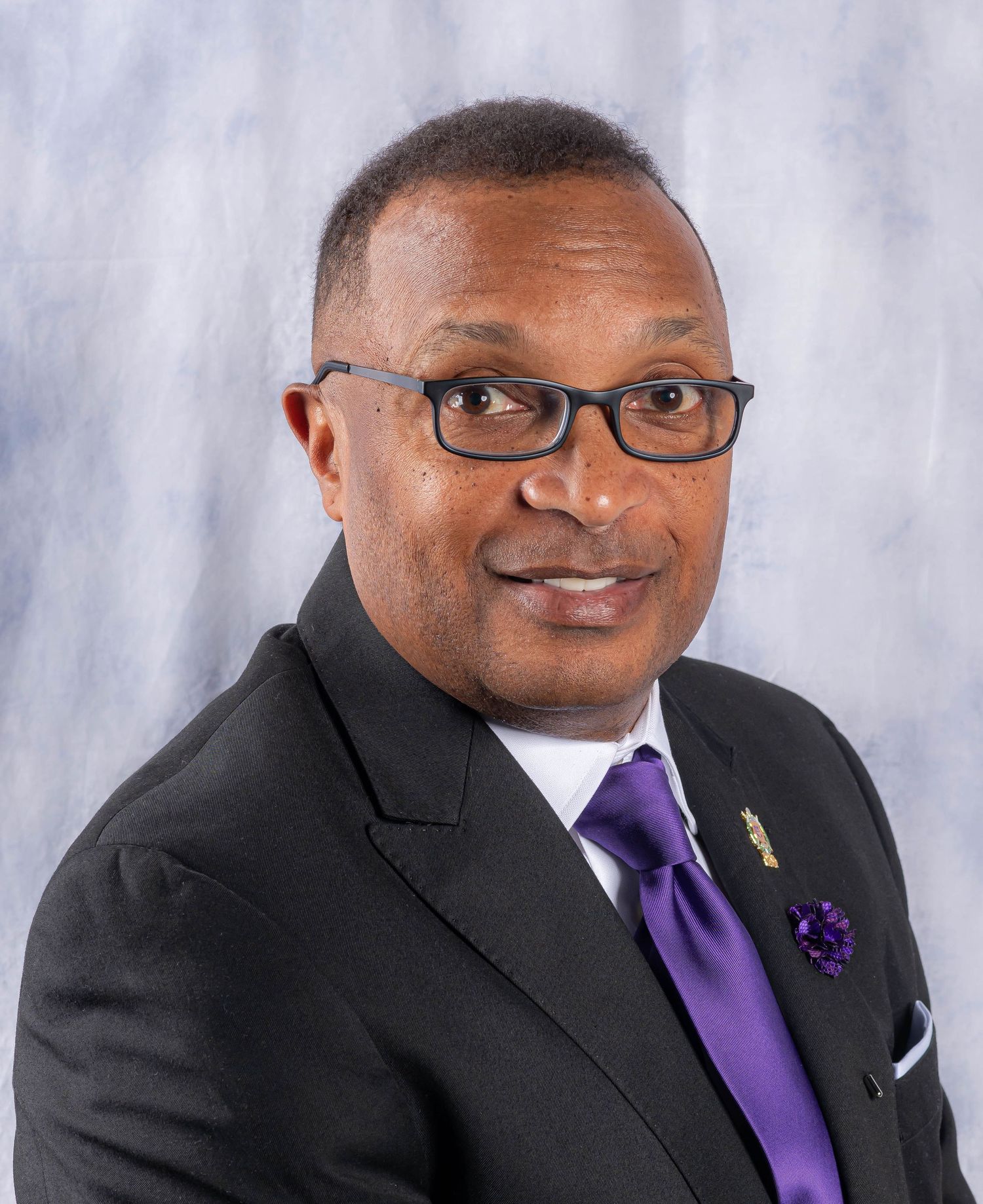 Professional headshot of person in black suit and purple tie against light backdrop.