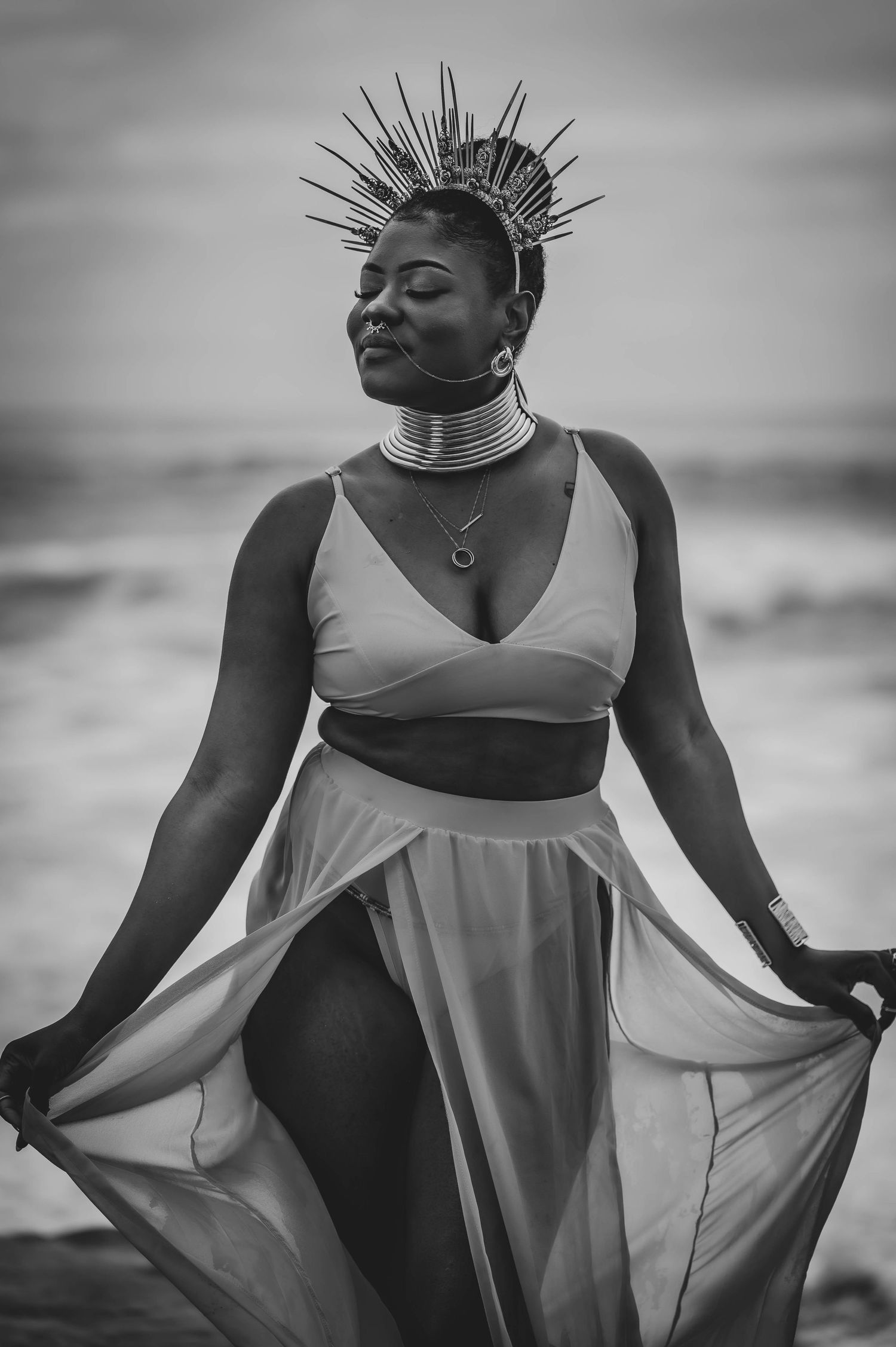 Person wearing spiked crown and neck rings poses in flowy dress against ocean backdrop.