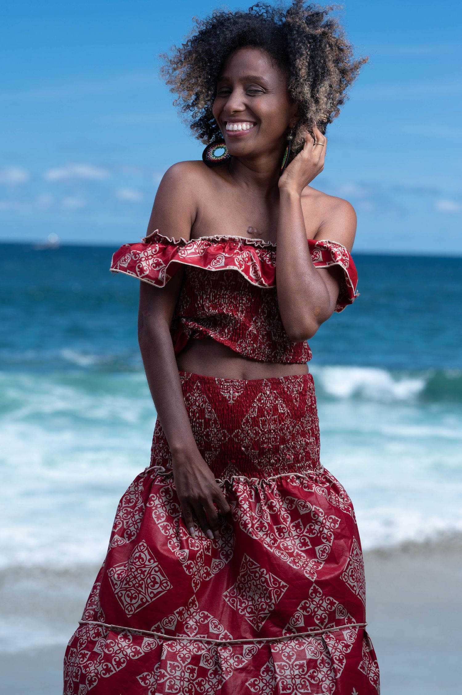 Woman in red and white patterned dress poses playfully on a beach with ocean waves in background.