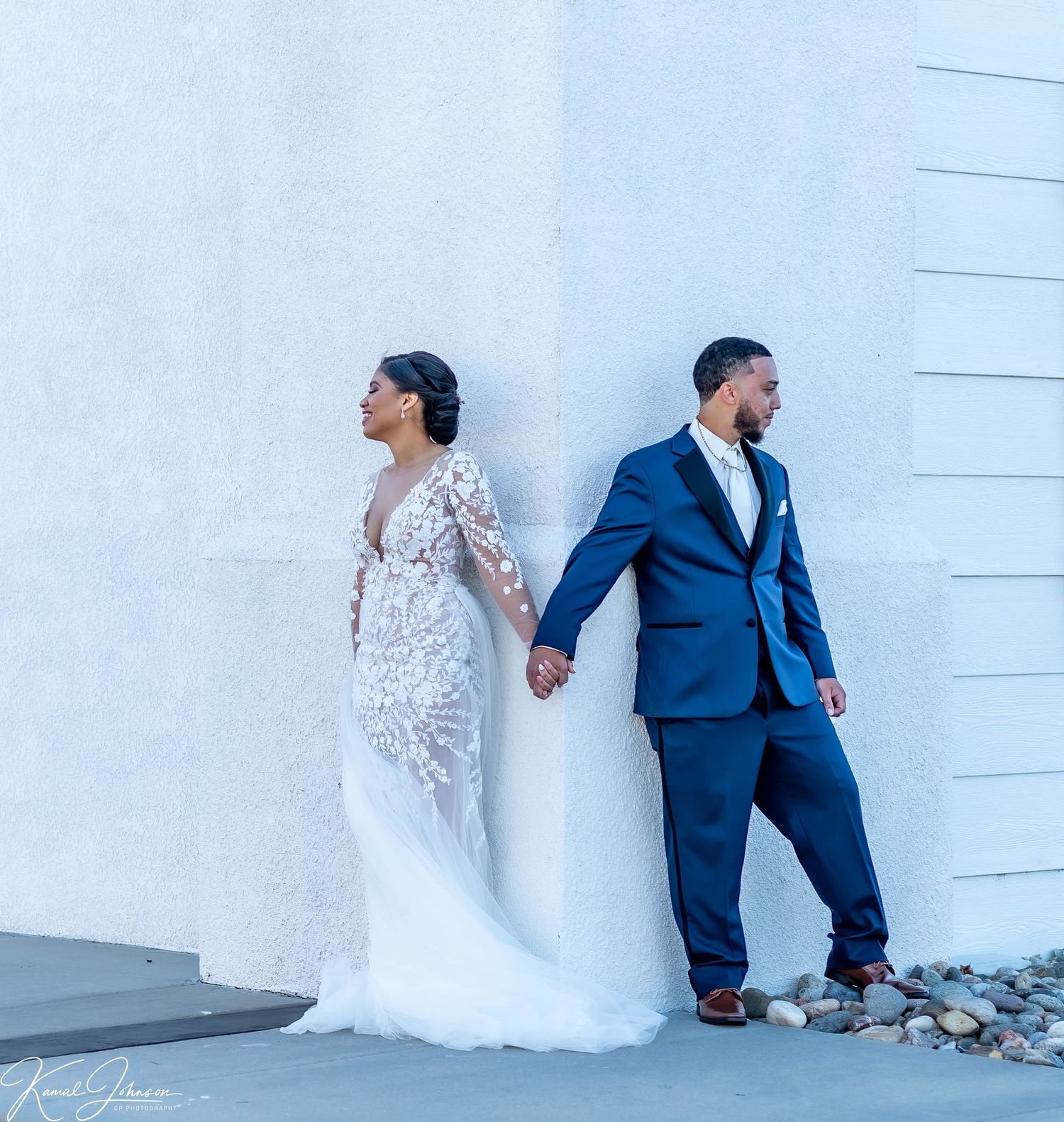 Couple in formal wedding attire poses against white wall.