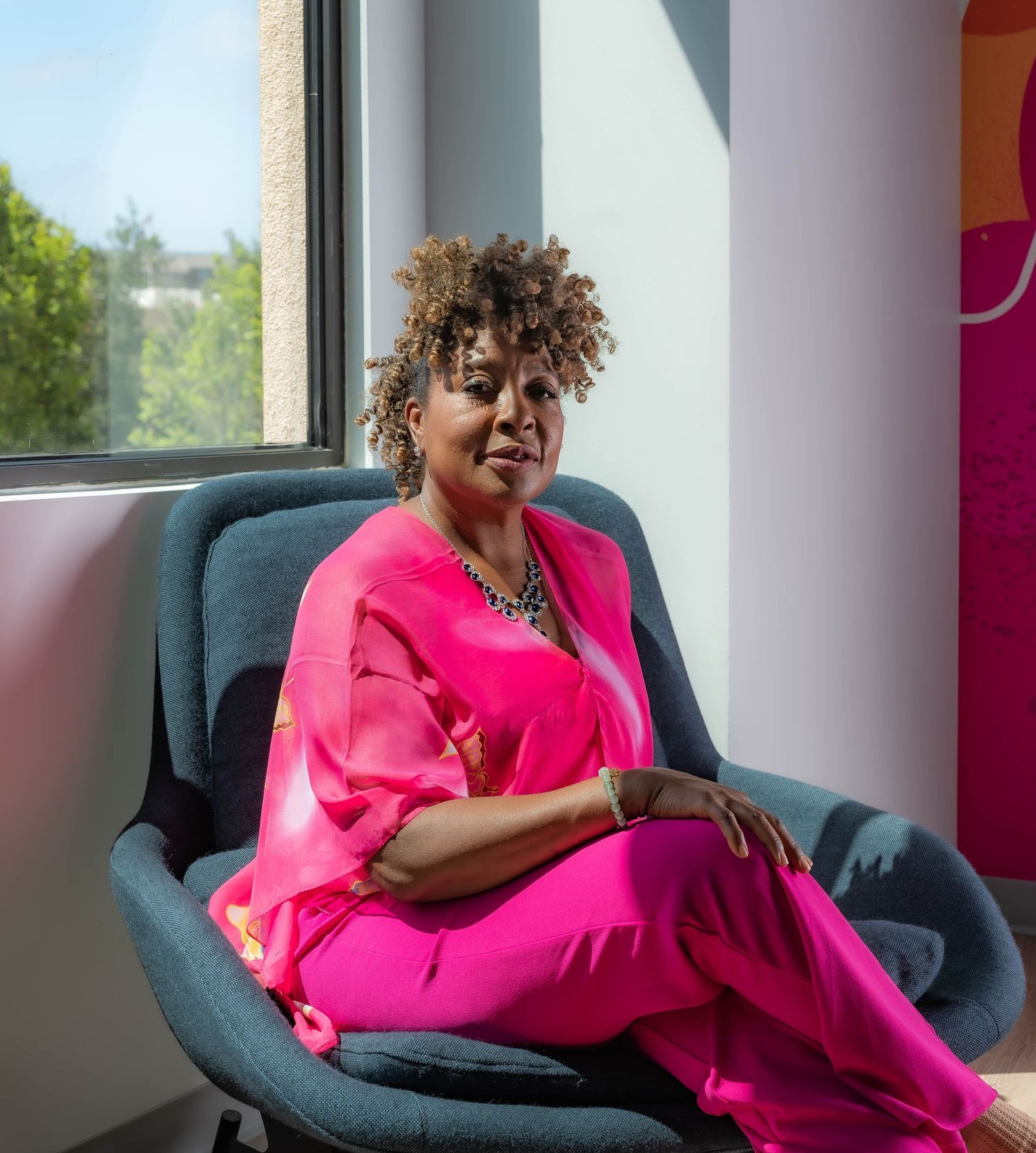 Person in bright pink blazer relaxes on teal armchair near window.