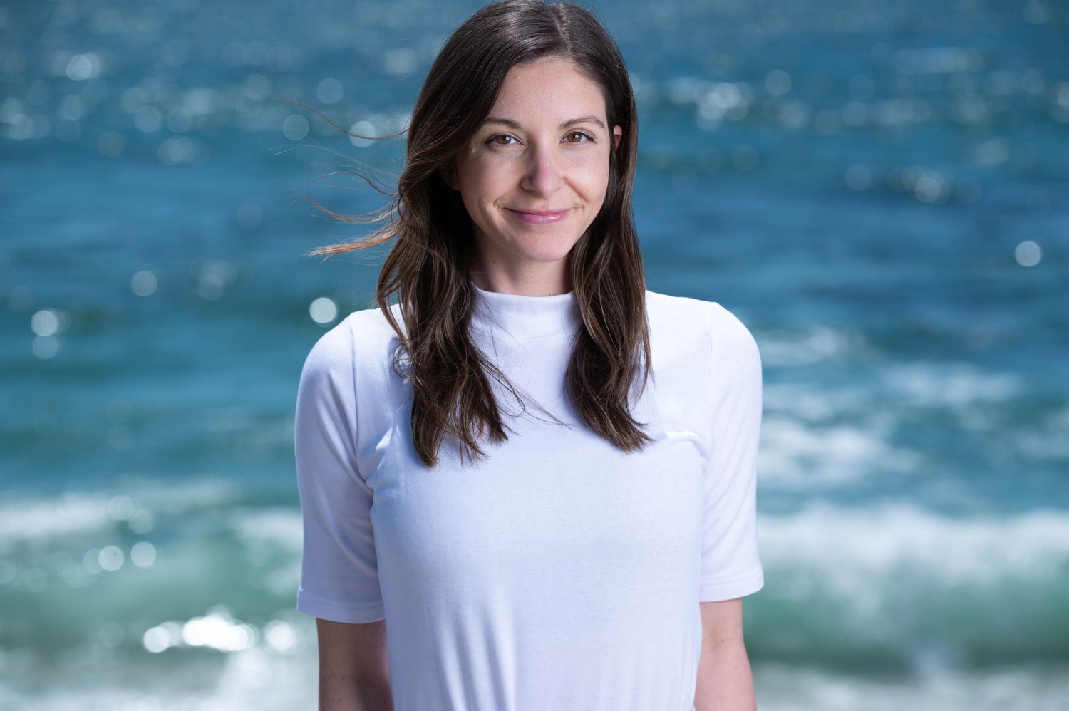Woman in white t-shirt smiling at beach with ocean waves and blue sky behind.