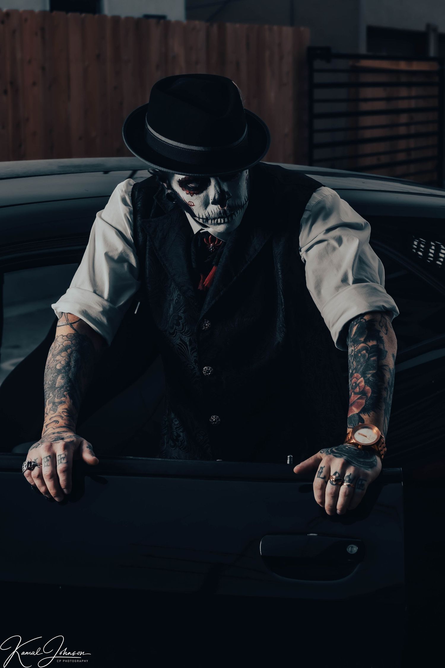 Person in formal wear with tattoos leaning against dark car.