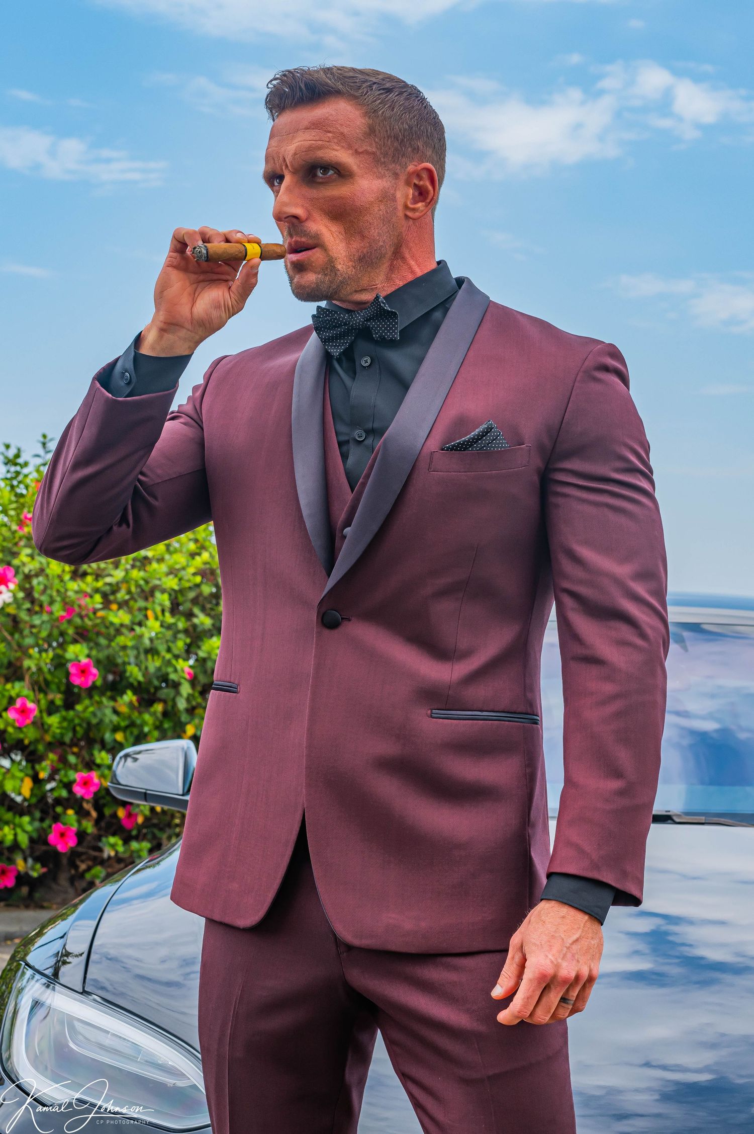 Man wearing burgundy suit and black tie posing against ocean backdrop.
