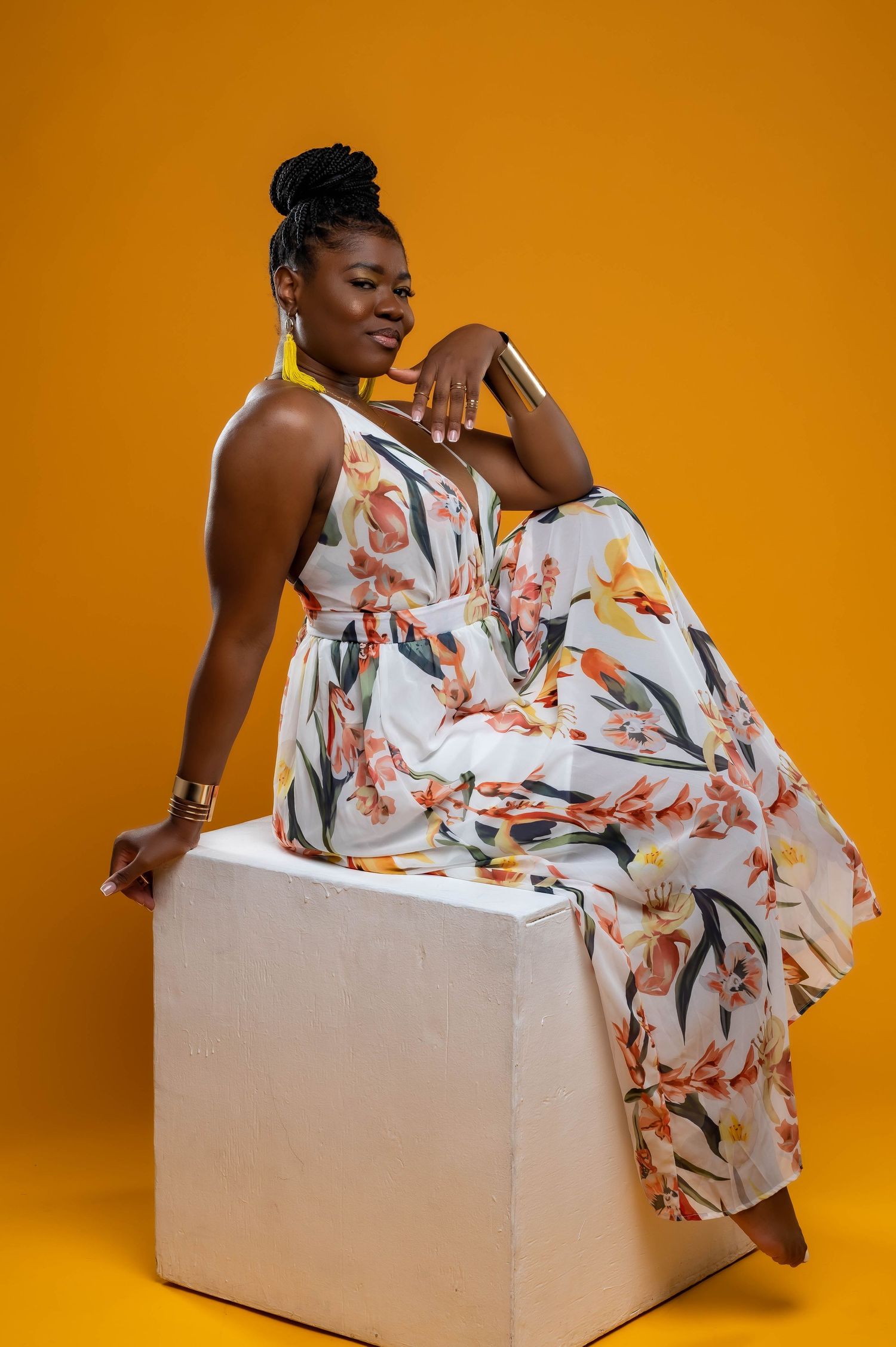 Model in floral maxi dress poses elegantly on white cube against vibrant orange background.