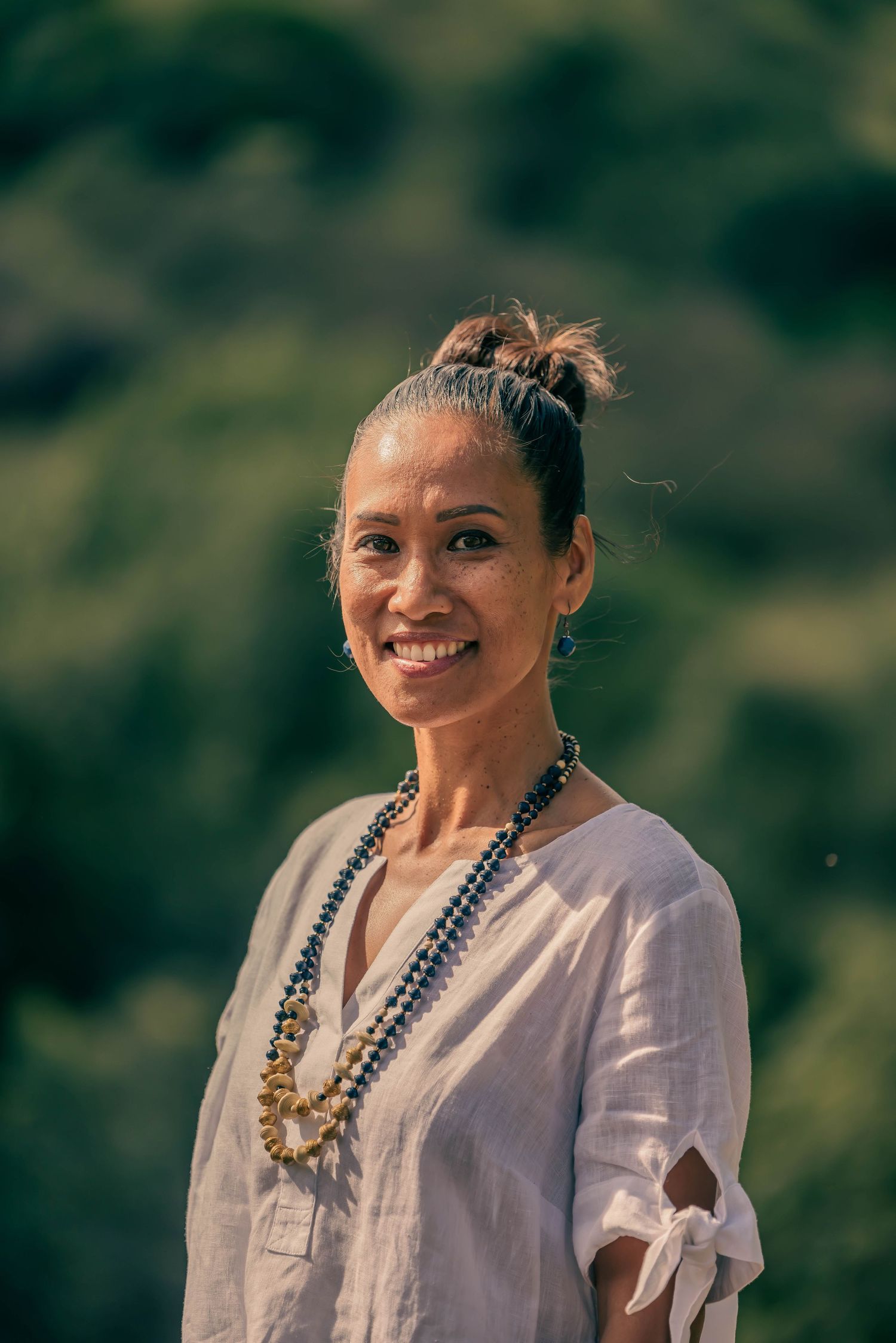 A person in a white blouse and statement necklace smiles warmly against a natural outdoor background.