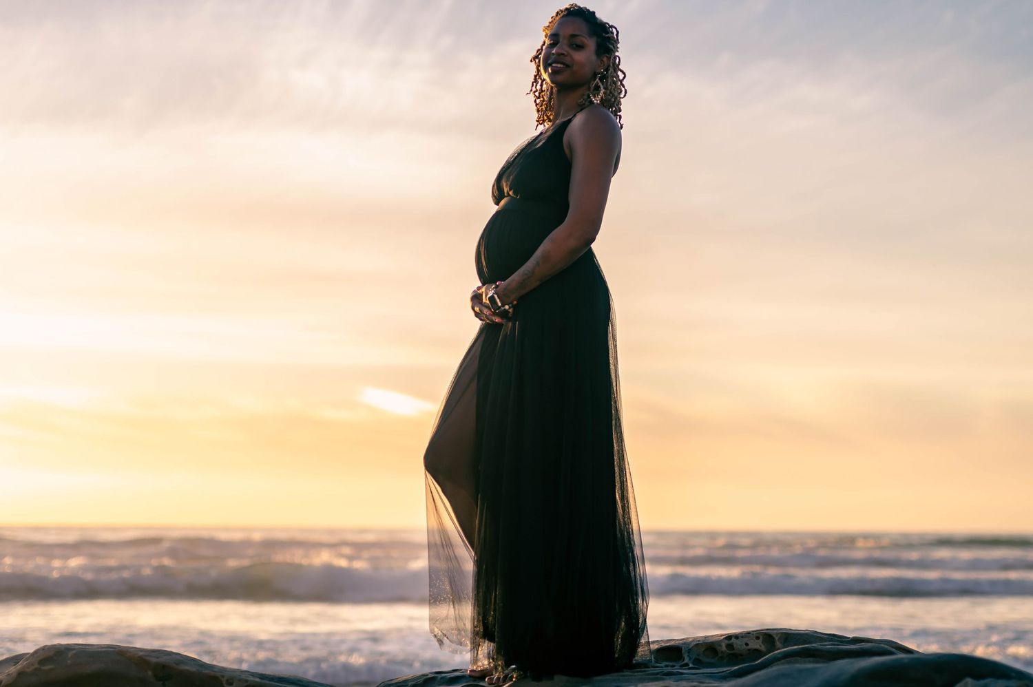 Silhouetted pregnant woman in flowing dress poses at beach during sunset.