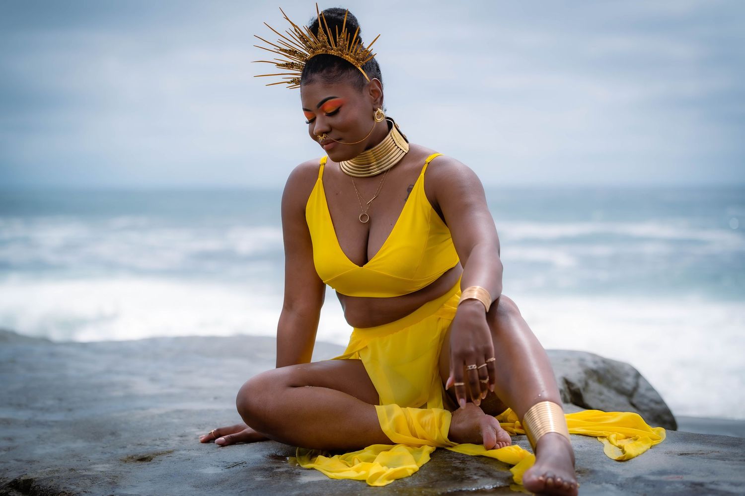 Woman in yellow dress poses gracefully on beach with cloudy ocean backdrop.