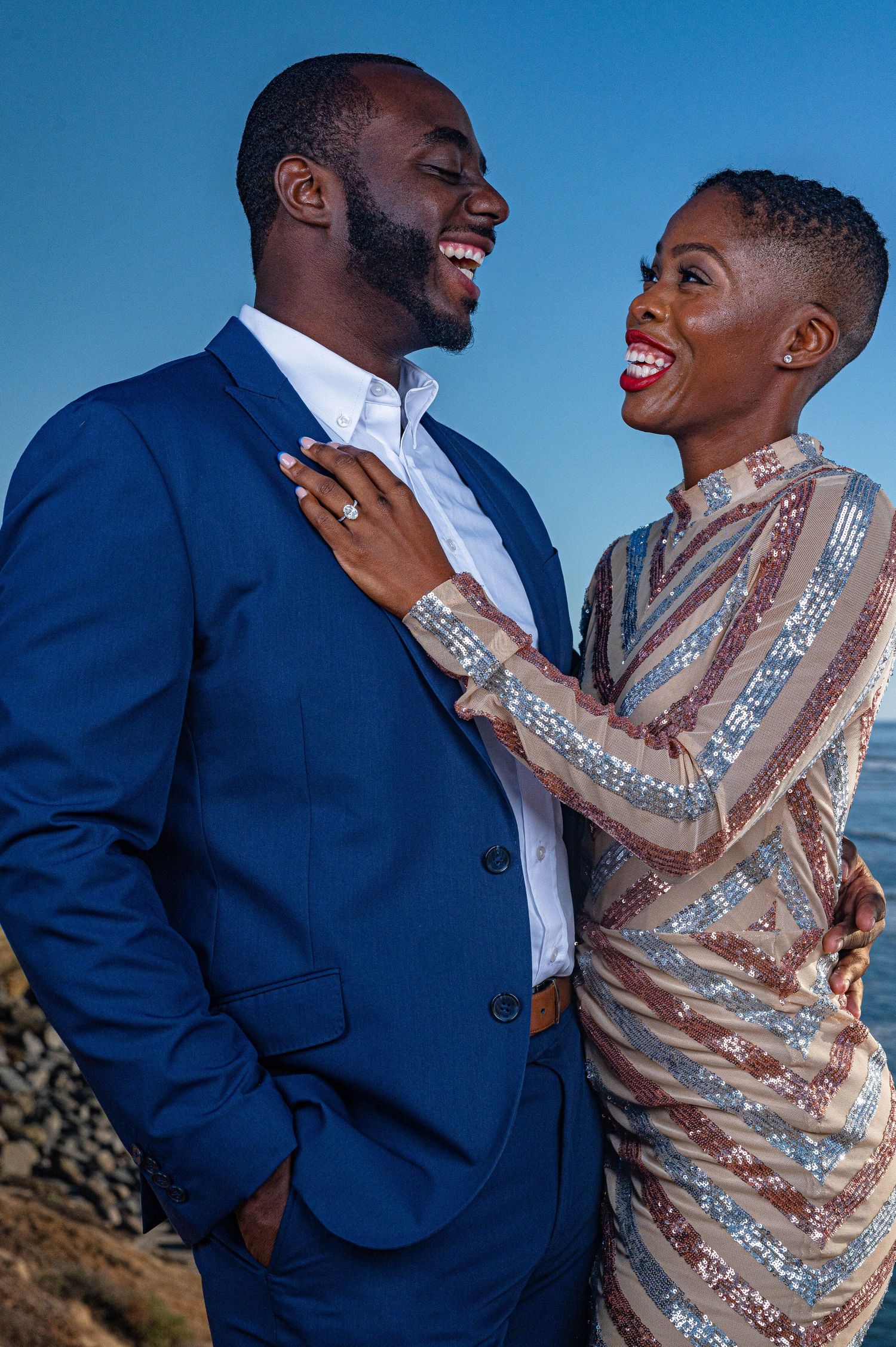 Engaged couple in formal wear sharing romantic moment against blue sky background.