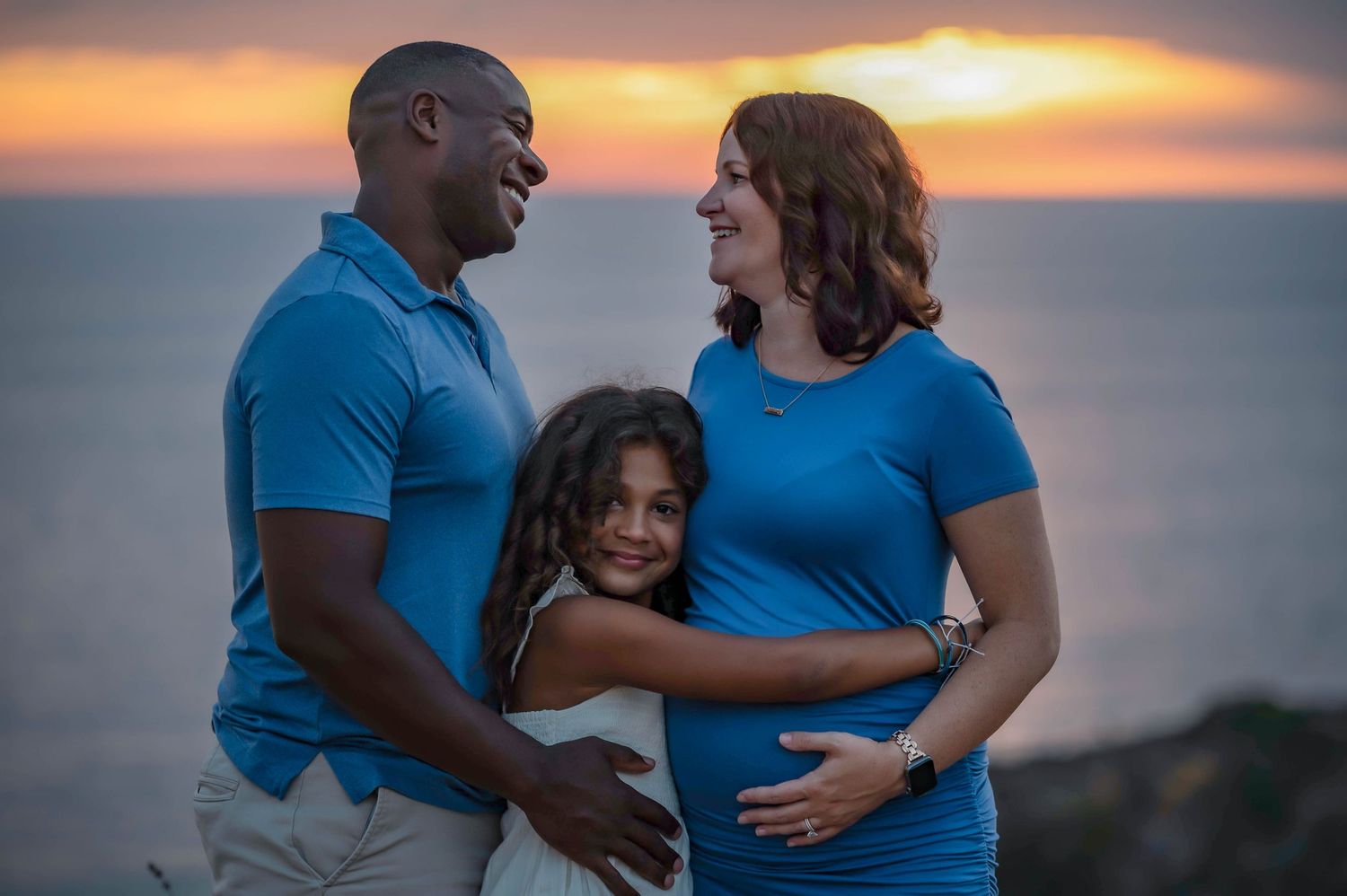 Family of three poses for maternity photo at sunset with ocean backdrop.