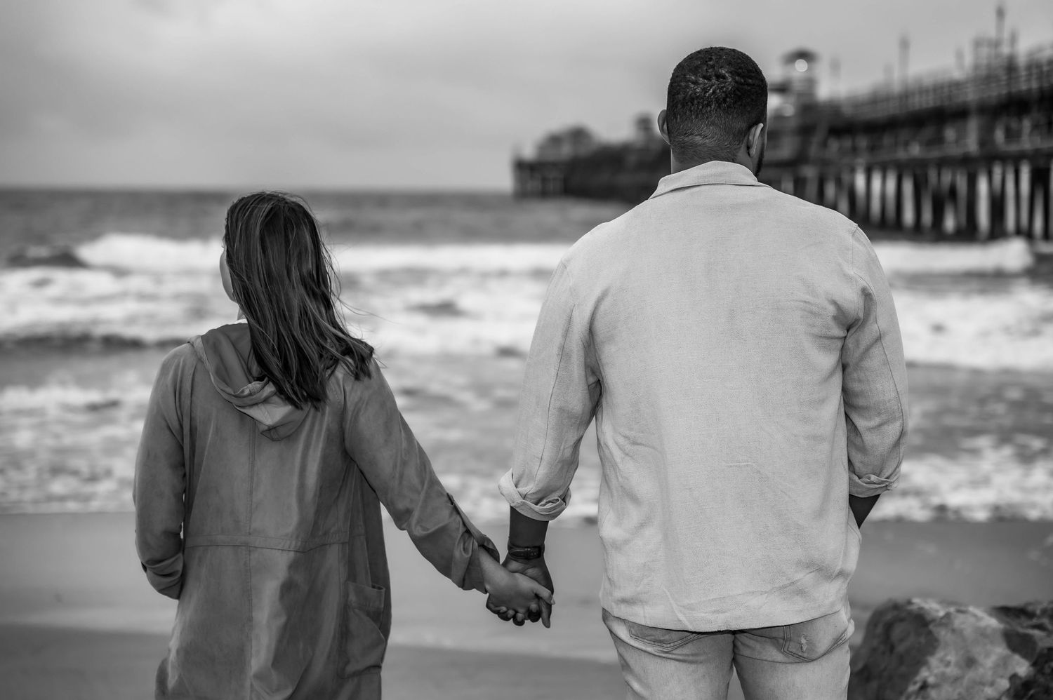 Couple holding hands while walking on beach near pier in black and white photography.