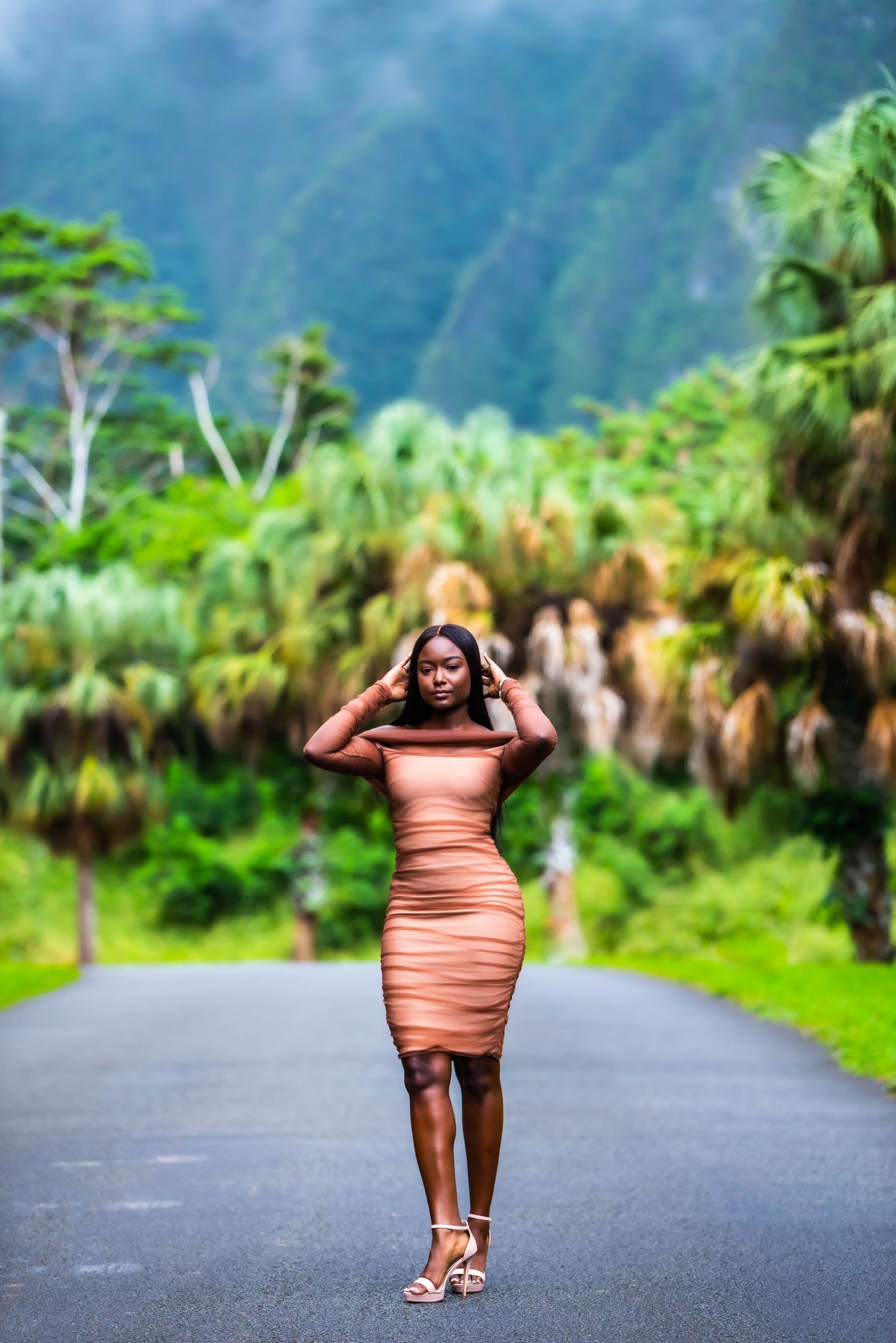 Person in coral dress poses playfully on tropical palm-lined road with mountains in background.