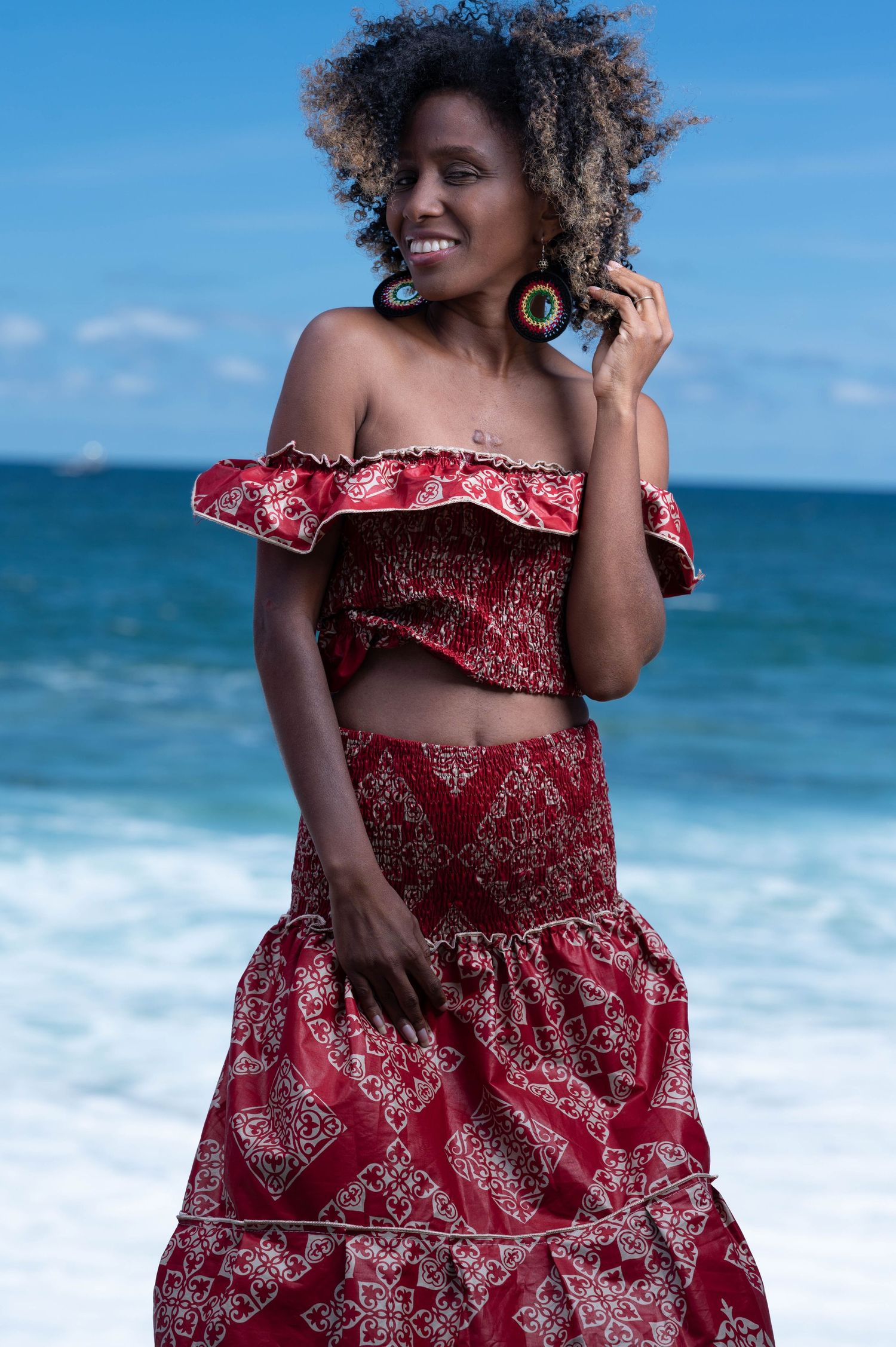 Woman in red patterned dress posing on beach with turquoise ocean waves behind her.