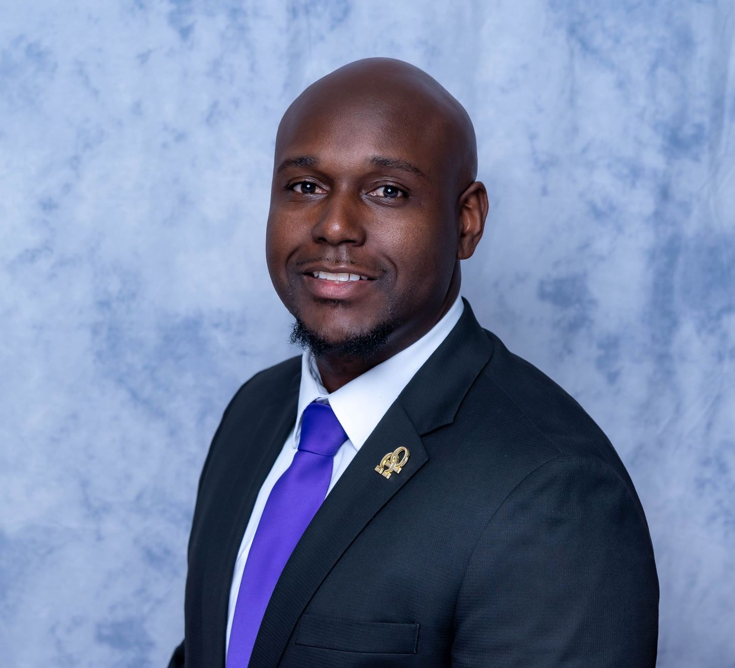 Professional portrait in black suit and purple tie against studio backdrop.