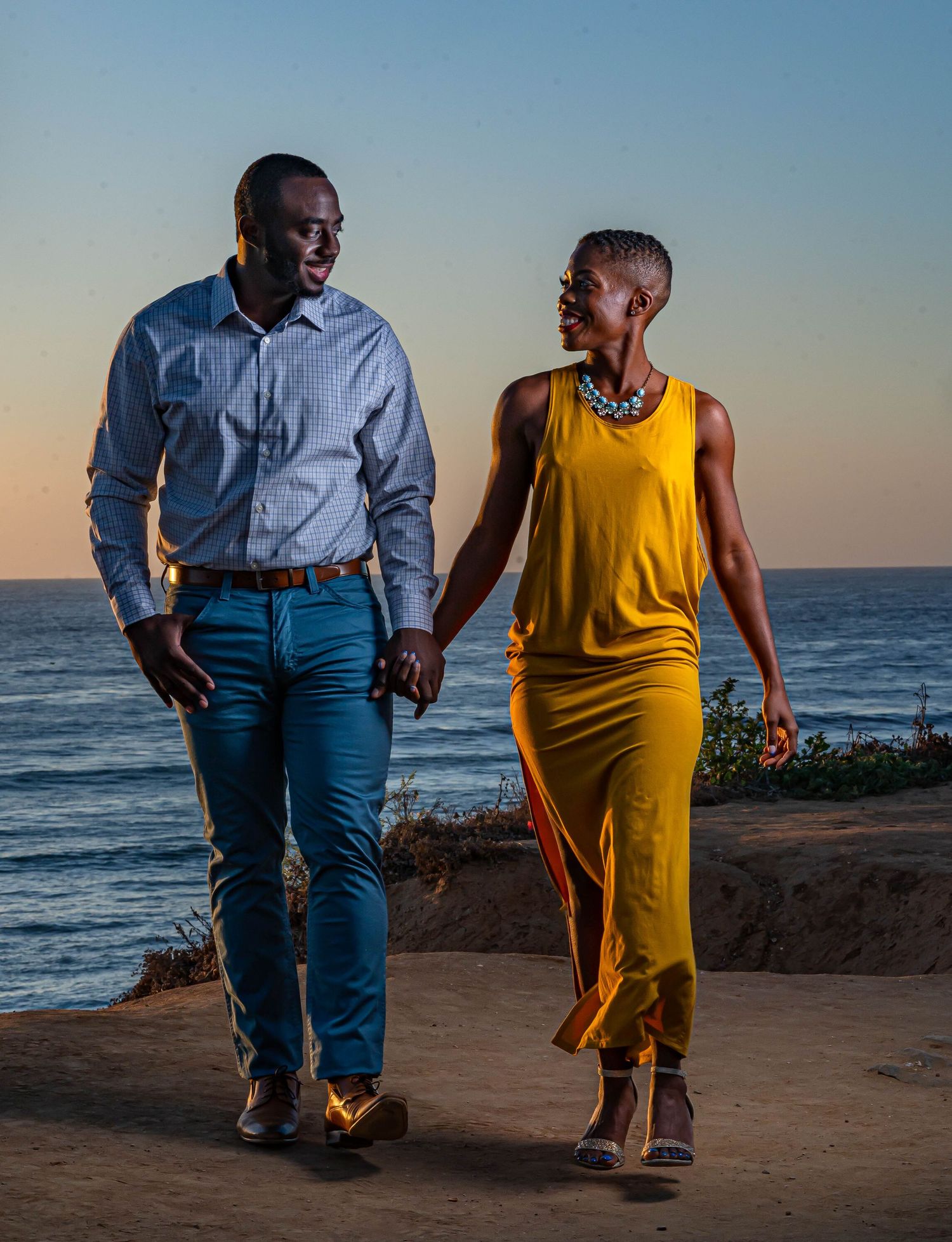 Couple walking hand in hand along ocean cliff at sunset in coordinated outfits.