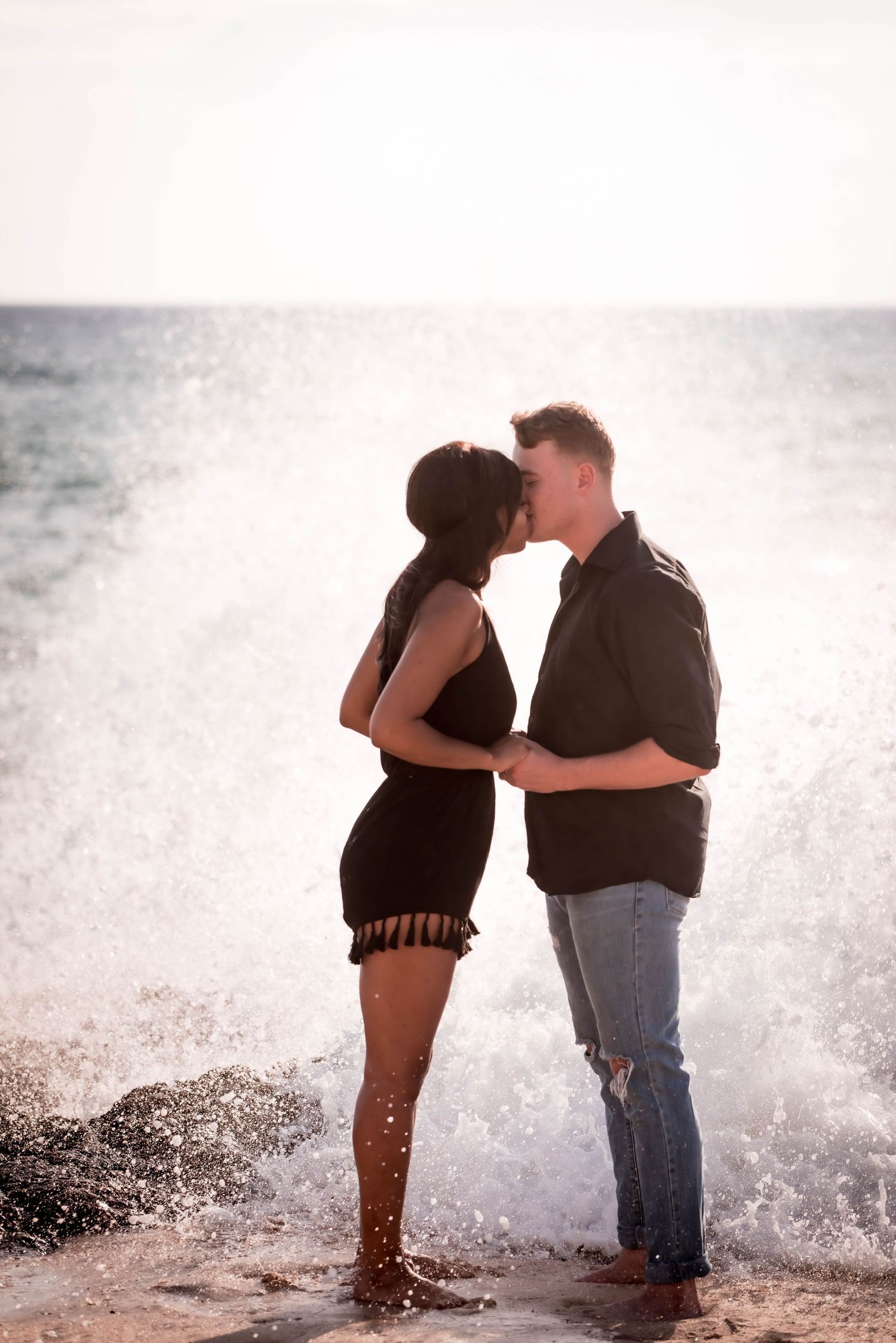 A romantic couple stands in silhouette against crashing ocean waves on a misty beach.
