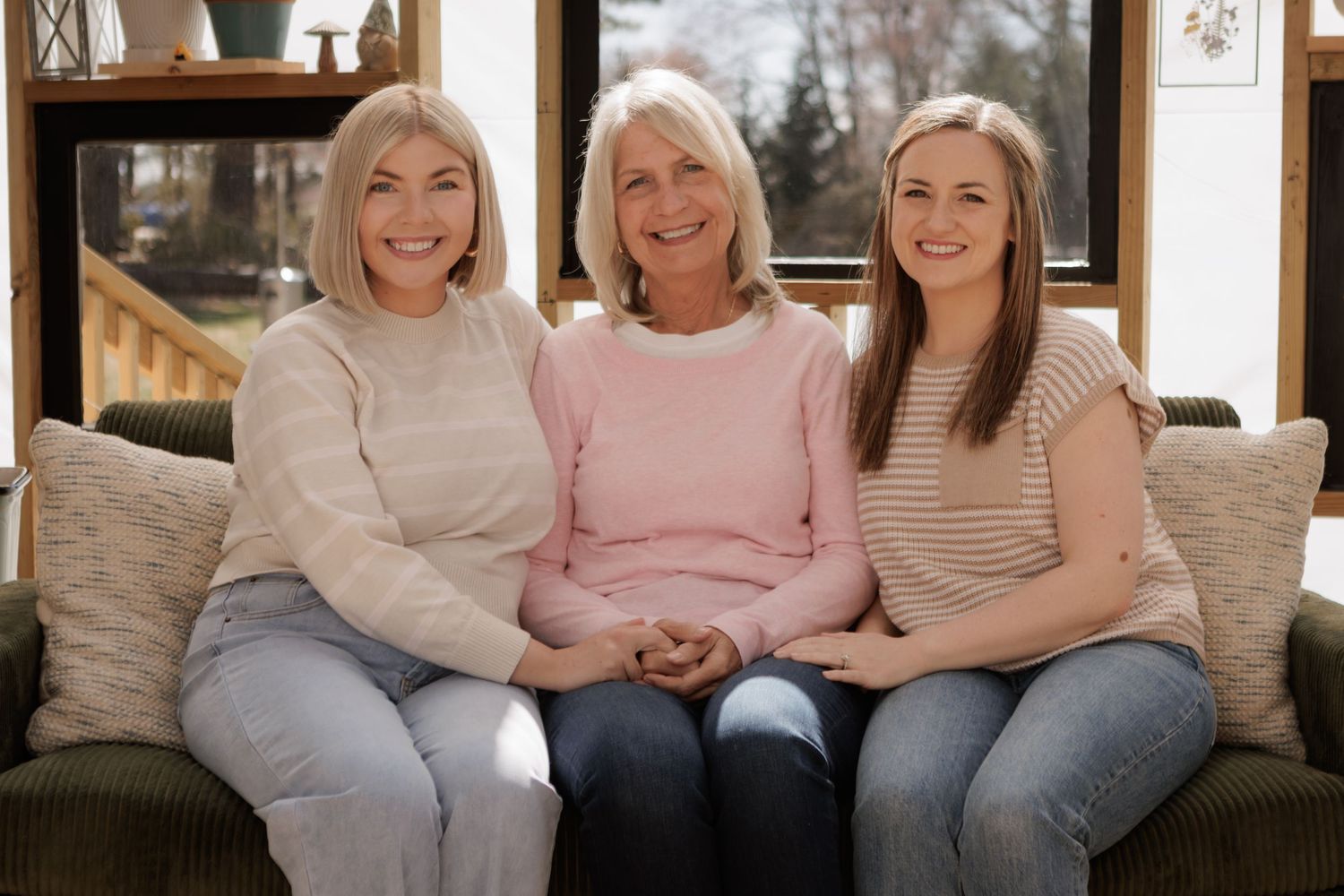 Three women of different generations smiling together on a couch in a bright living room.