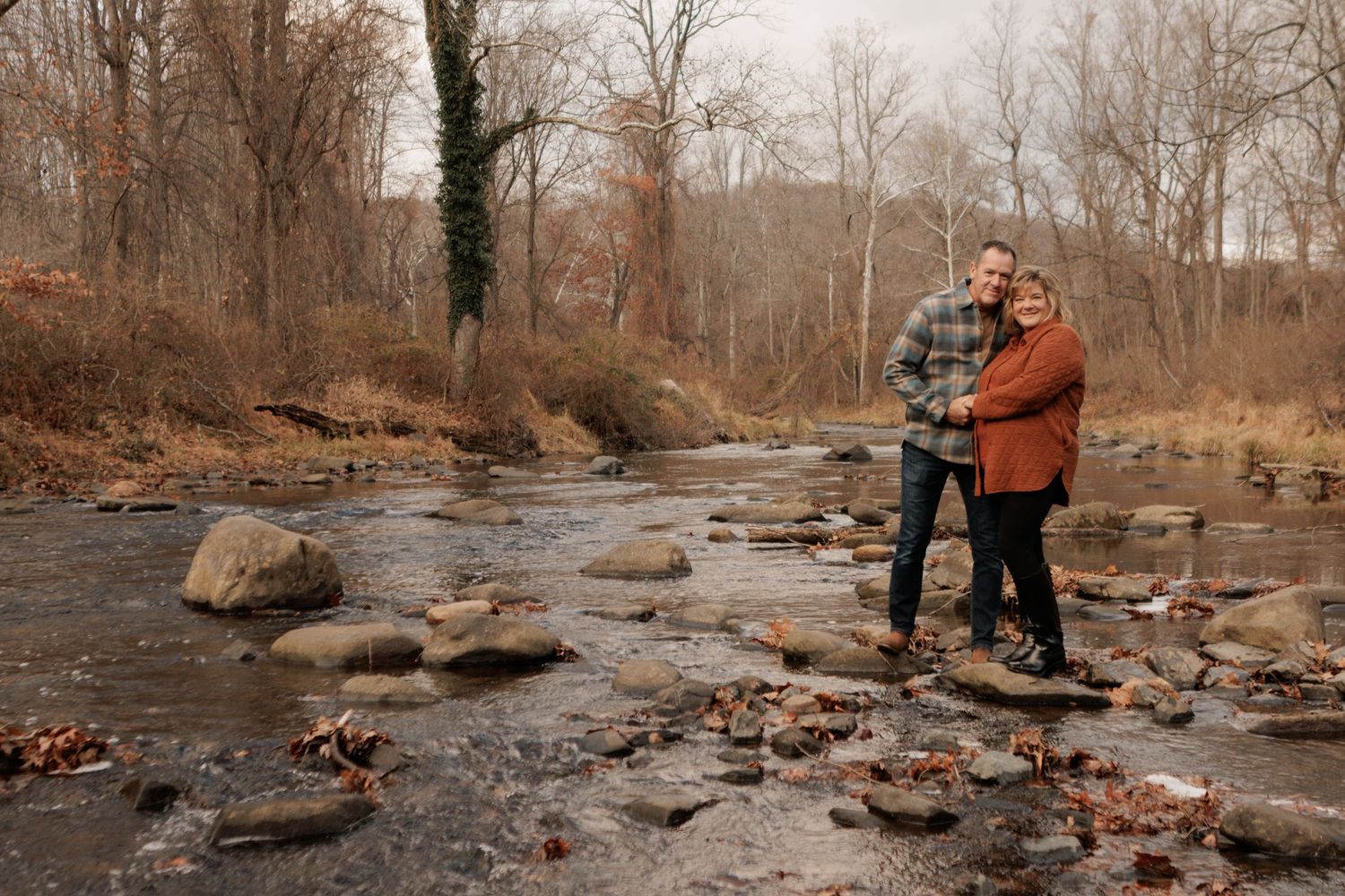 Couple embracing on rocks by a serene creek surrounded by bare autumn trees in a peaceful woodland setting.