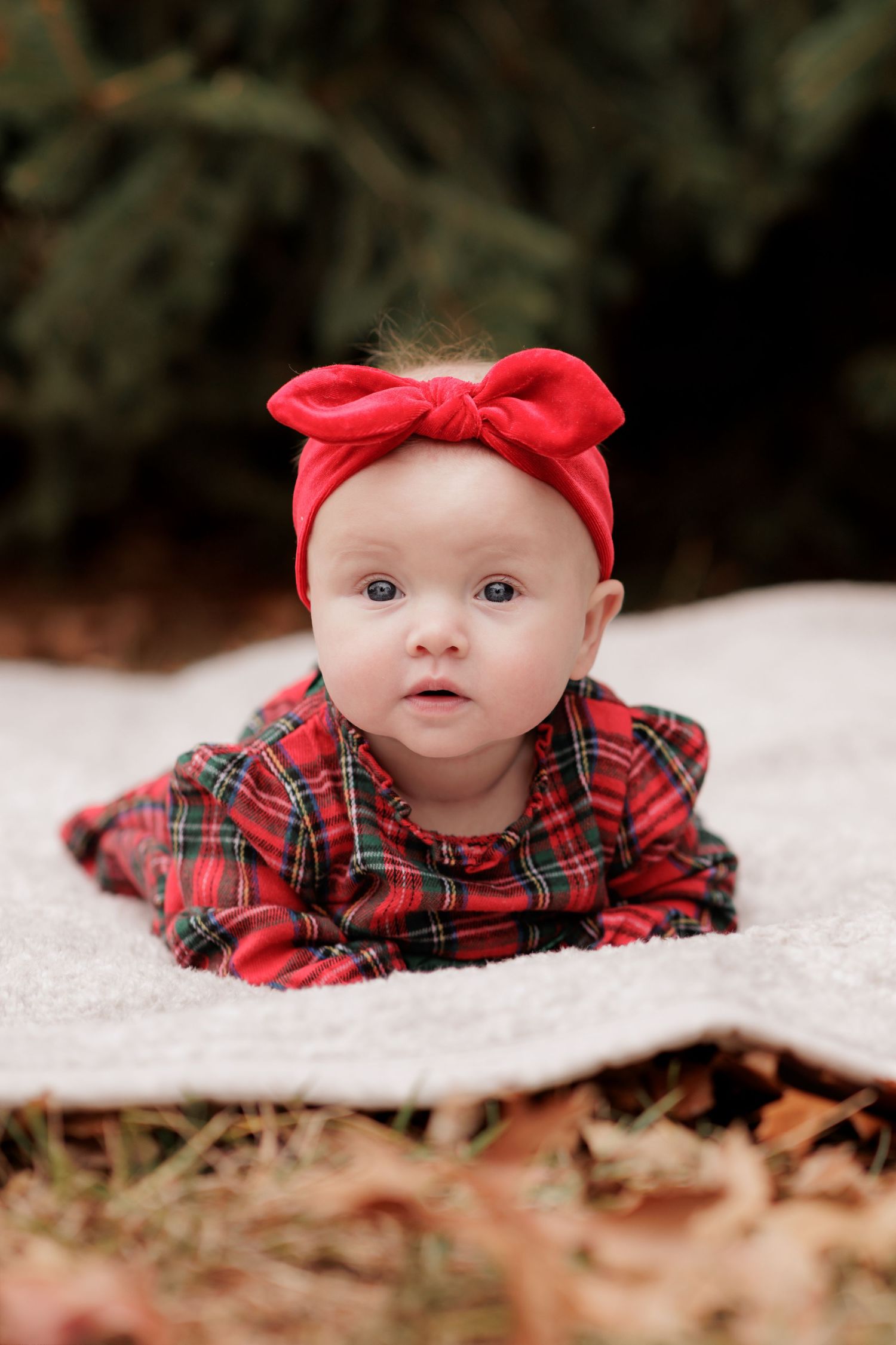 A baby wearing a red plaid outfit and bow headband lies on a white blanket.