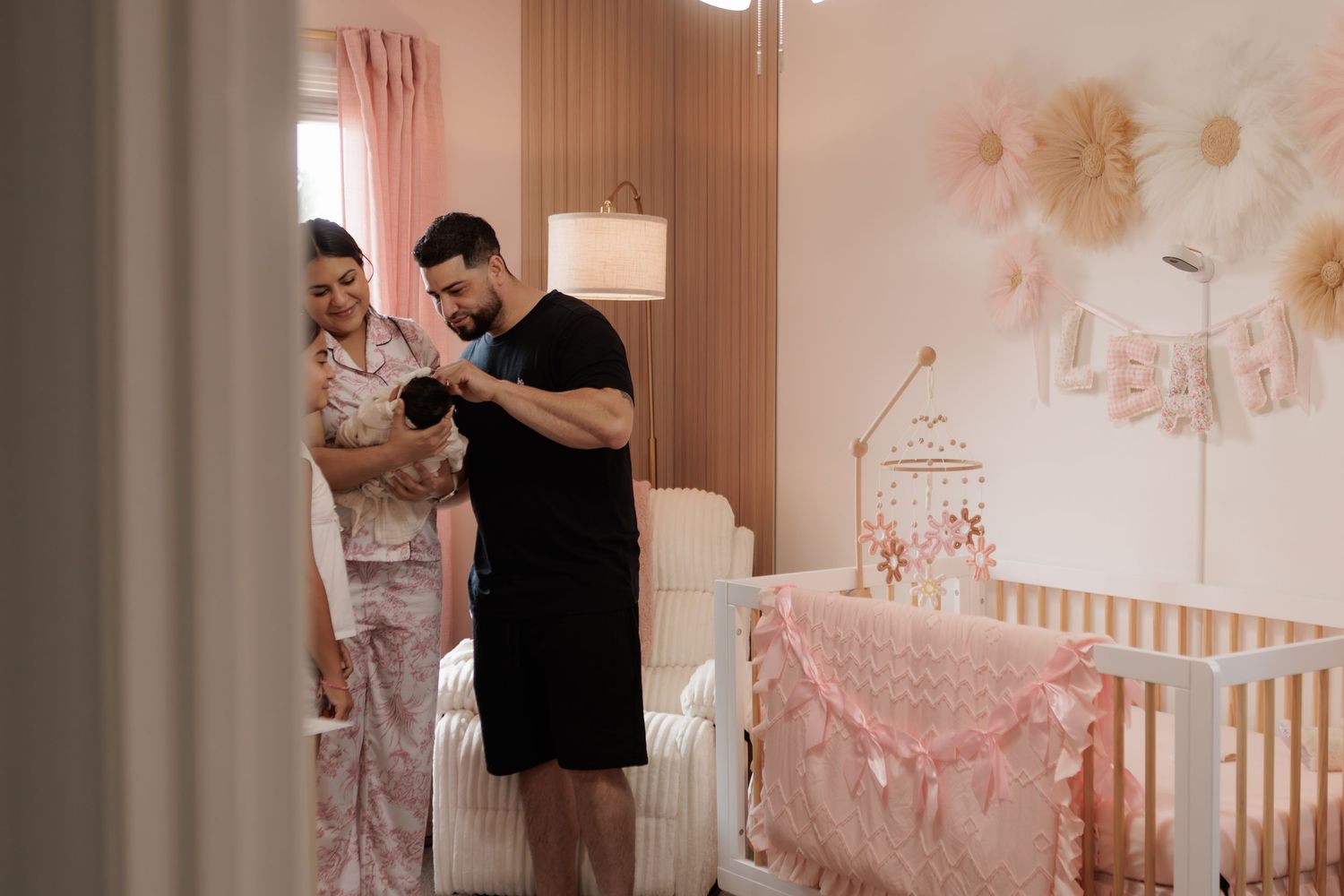 Parents standing together holding their newborn baby in a bright nursery with a white crib, pink blanket, and floral wall decor.
