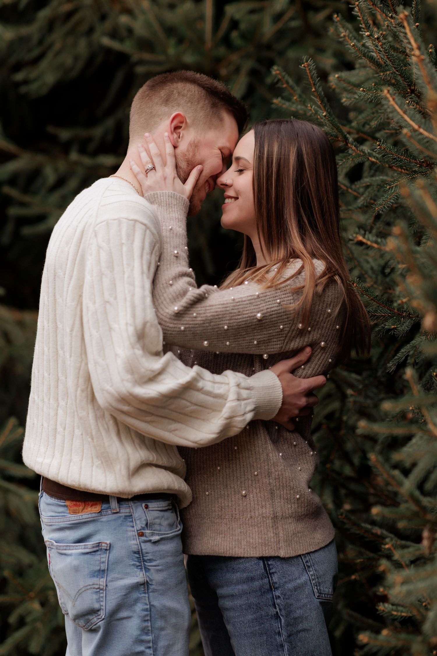 A couple in casual sweaters shares an intimate moment among evergreen trees.
