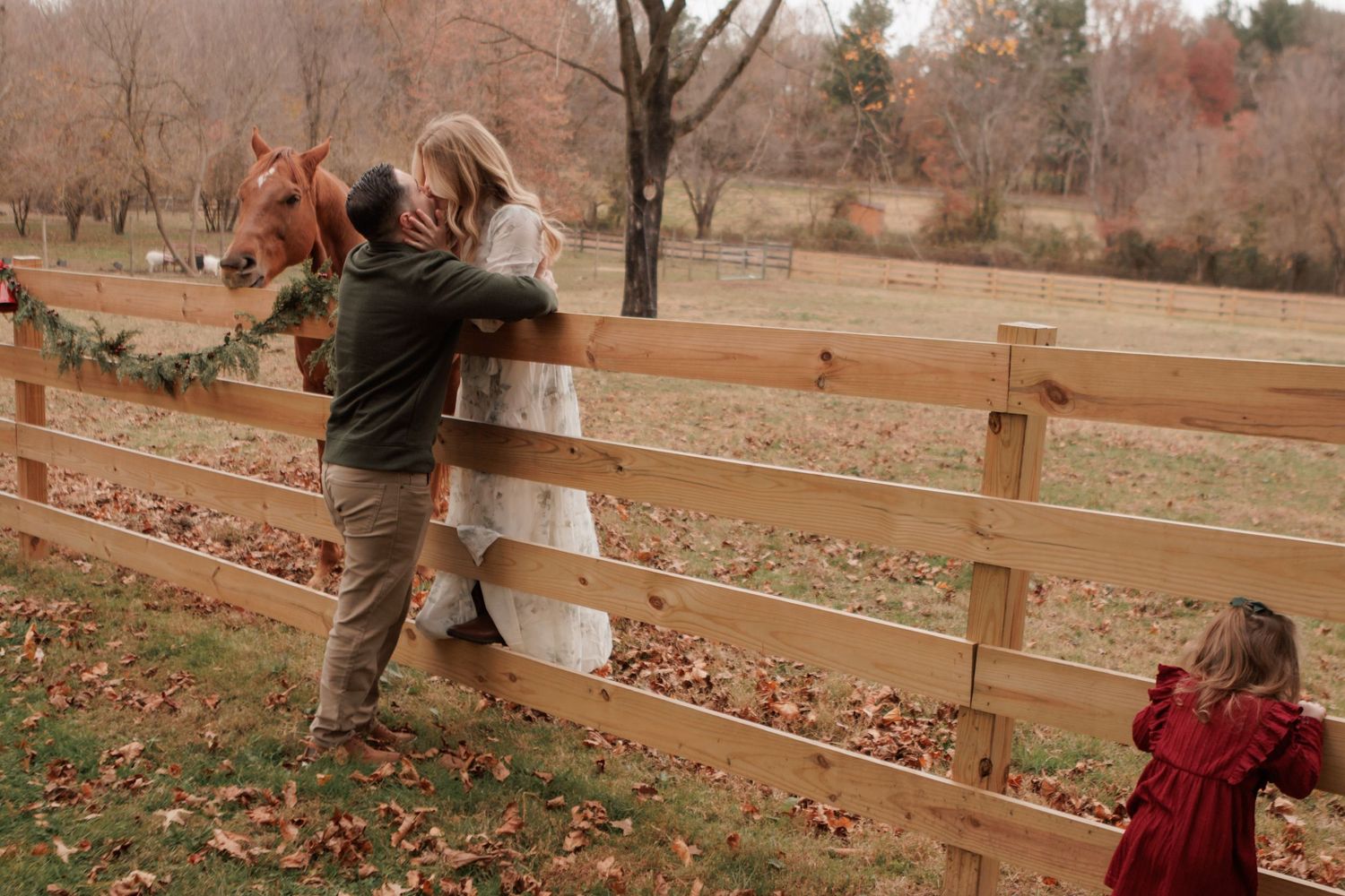 A horse watches over a wooden fence as people gather in an autumn farm setting.