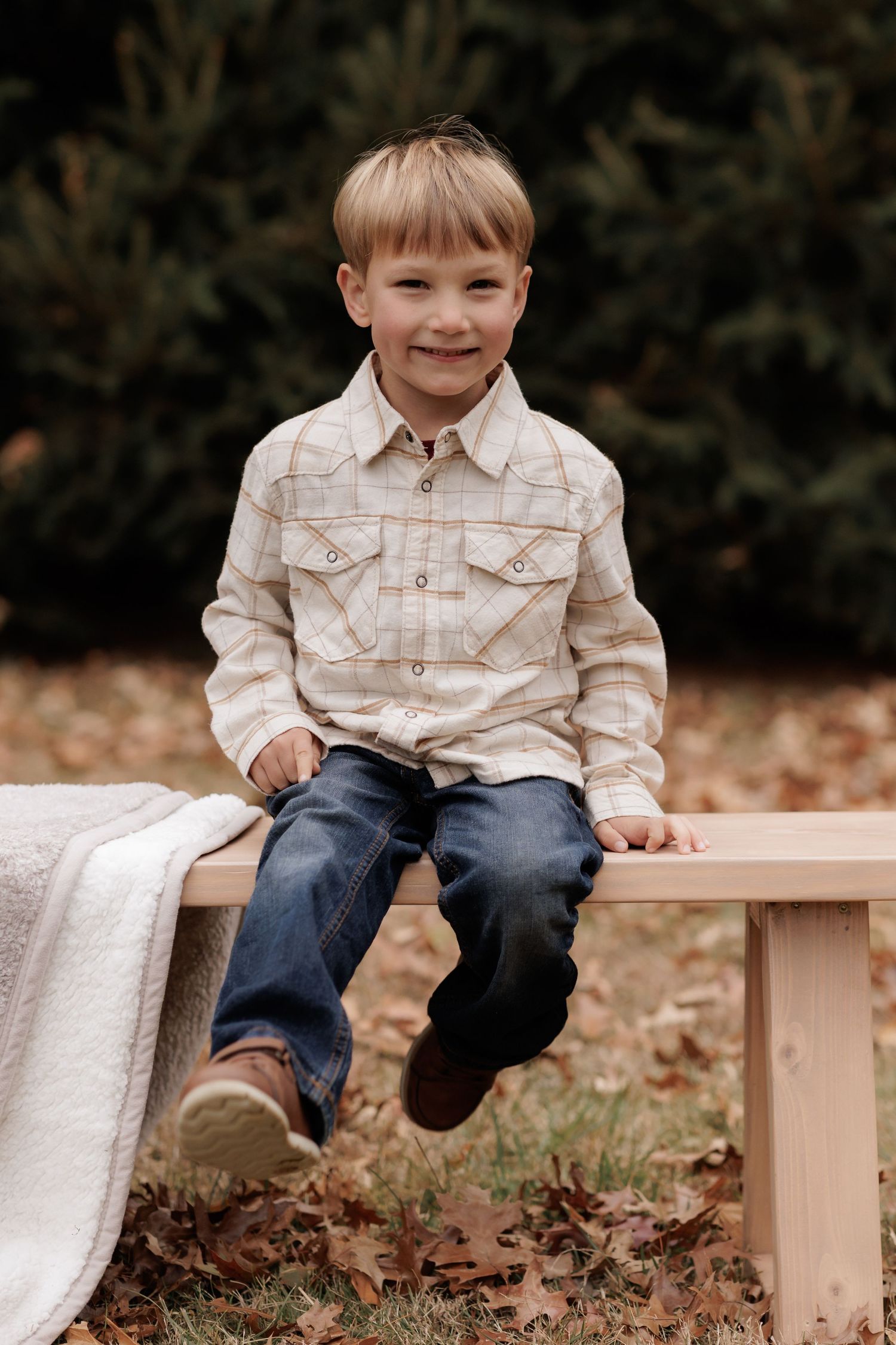 A child in a plaid shirt and jeans sits on a wooden bench surrounded by fallen autumn leaves in a garden setting.