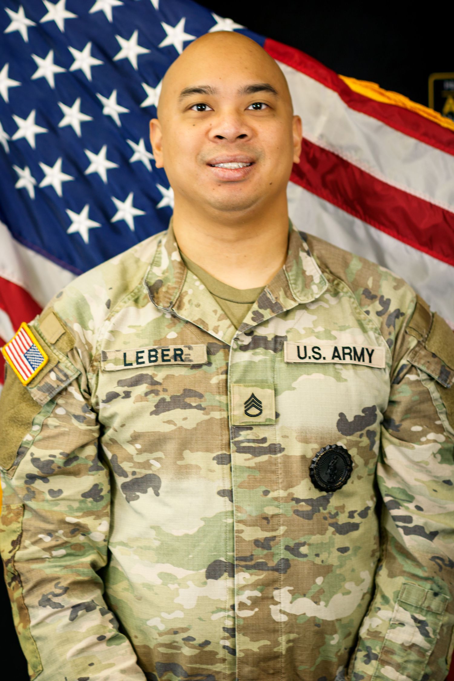 U.S. Army soldier in camouflage uniform poses in front of American flag.