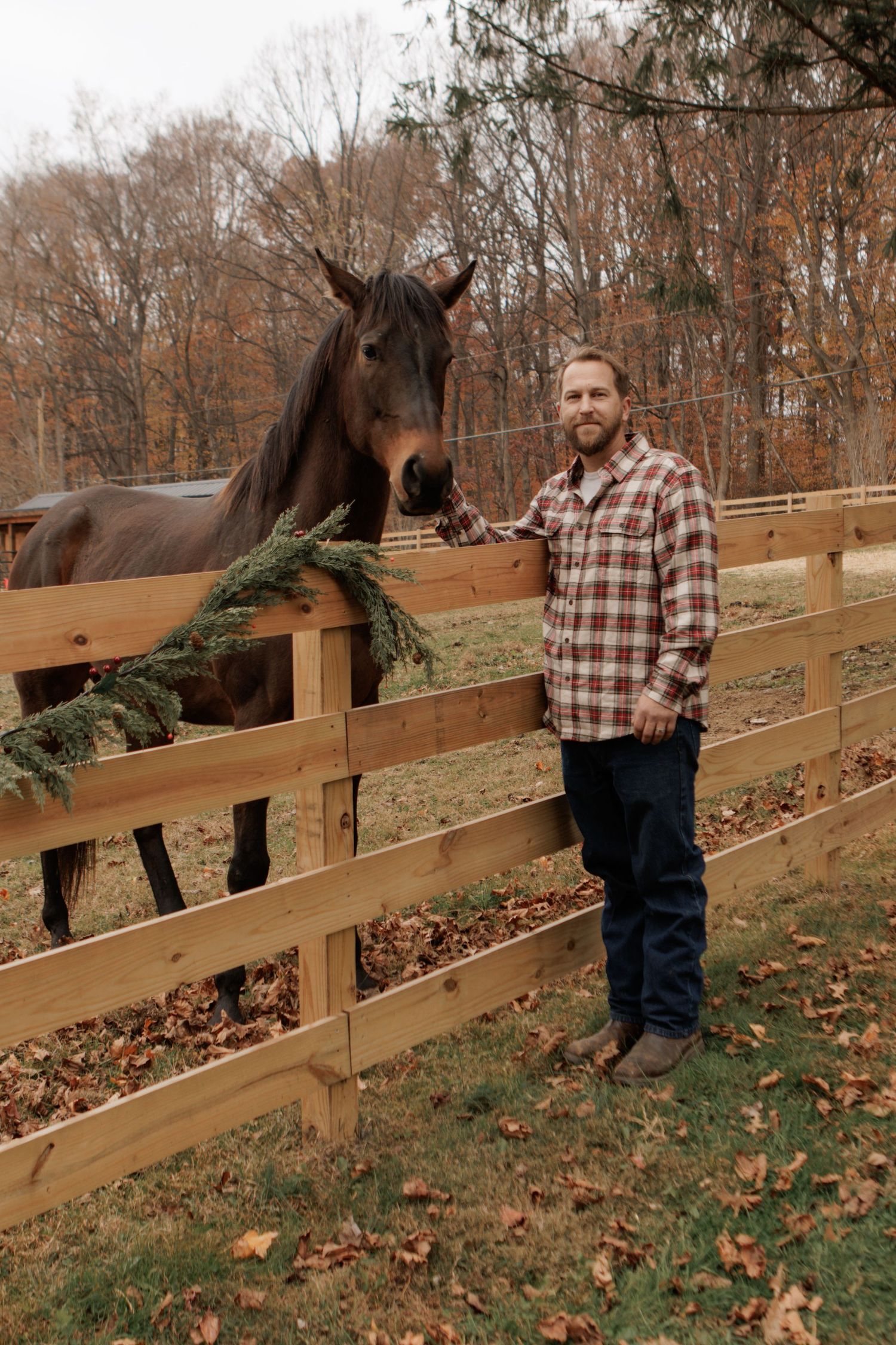 Brown horse and person in plaid shirt stand by wooden fence in autumn setting.