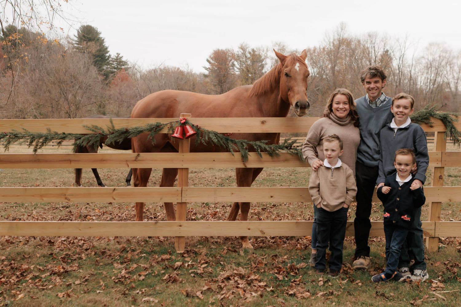 Family of five posing with a chestnut horse at a wooden fence decorated with holiday garland outdoors.