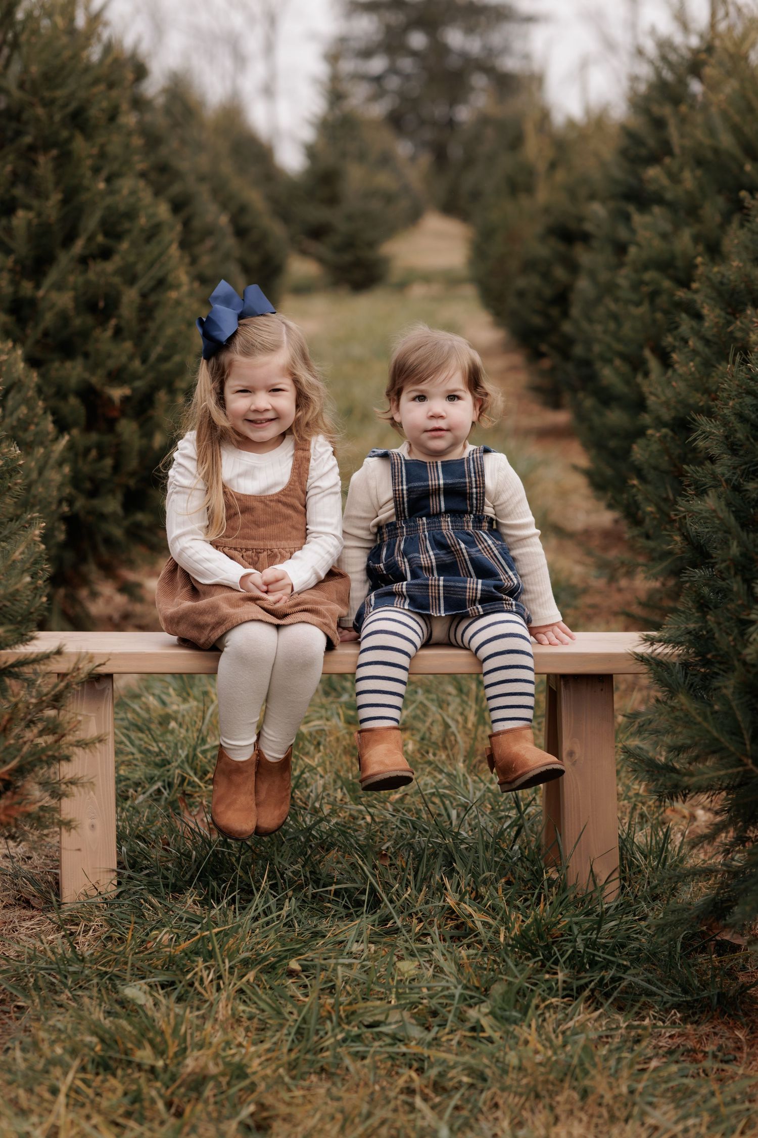 Two young girls sitting on a wooden bench at a Christmas tree farm, wearing festive outfits and boots.