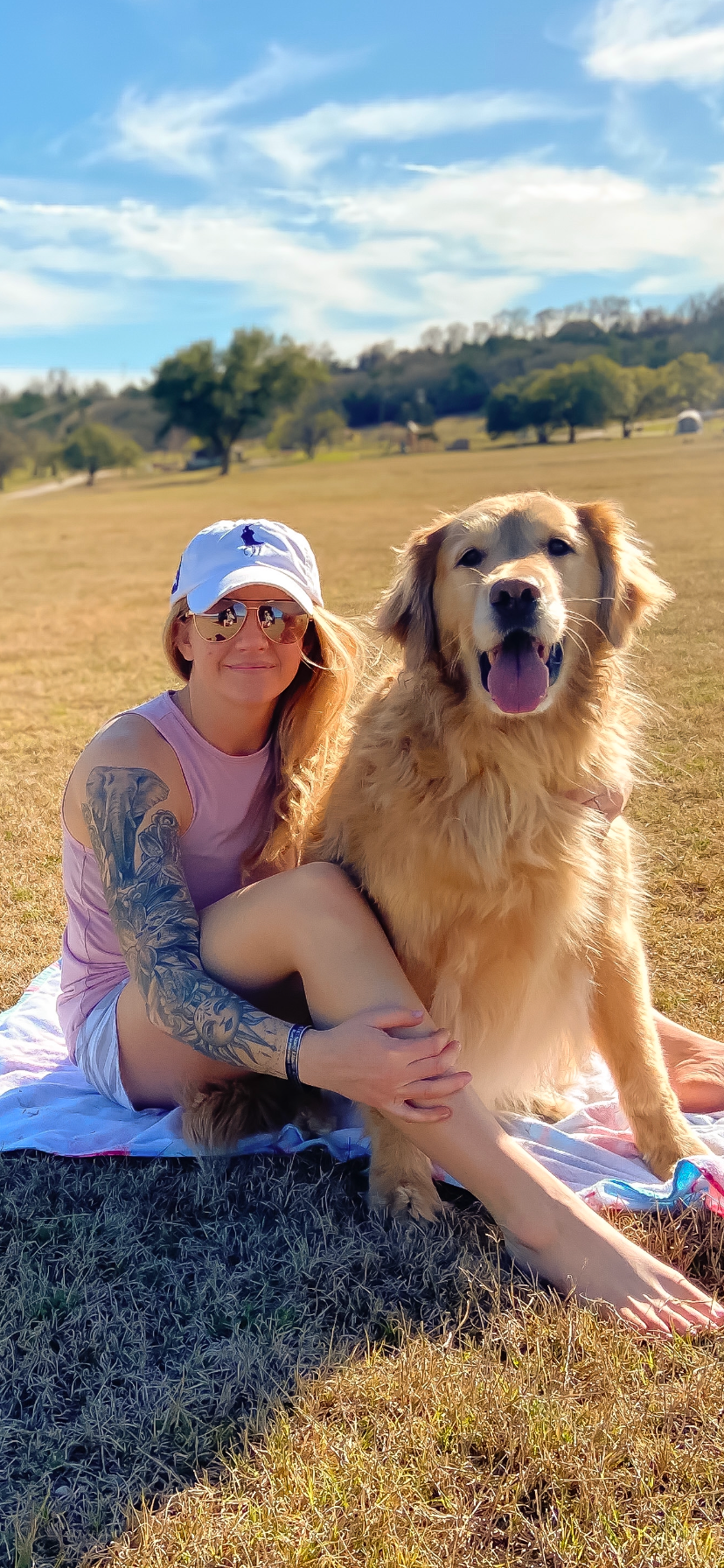 A happy Golden Retriever sits with someone on a grassy hill under blue skies.