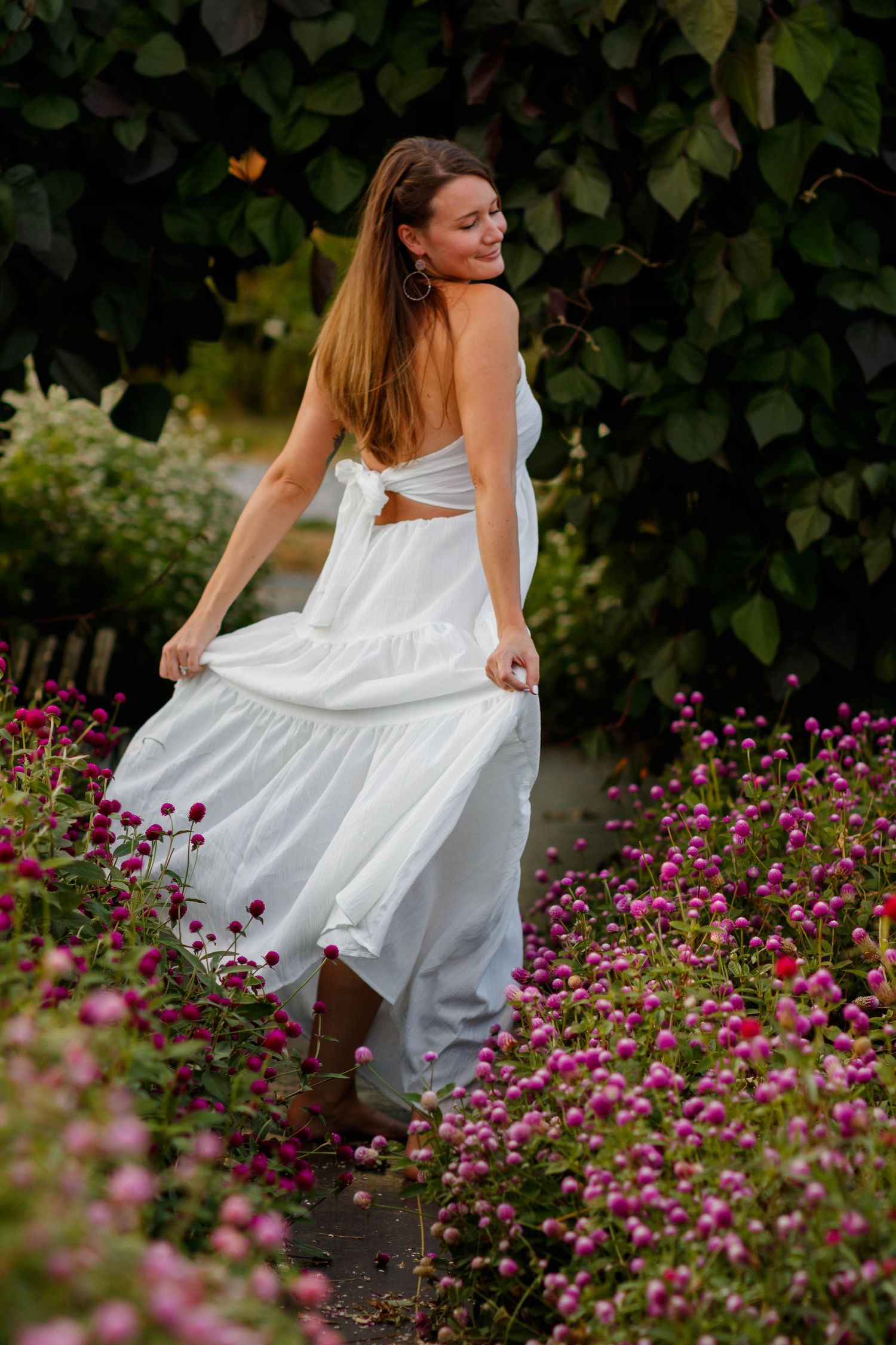Person in flowing white dress twirling among pink flowers in garden setting.