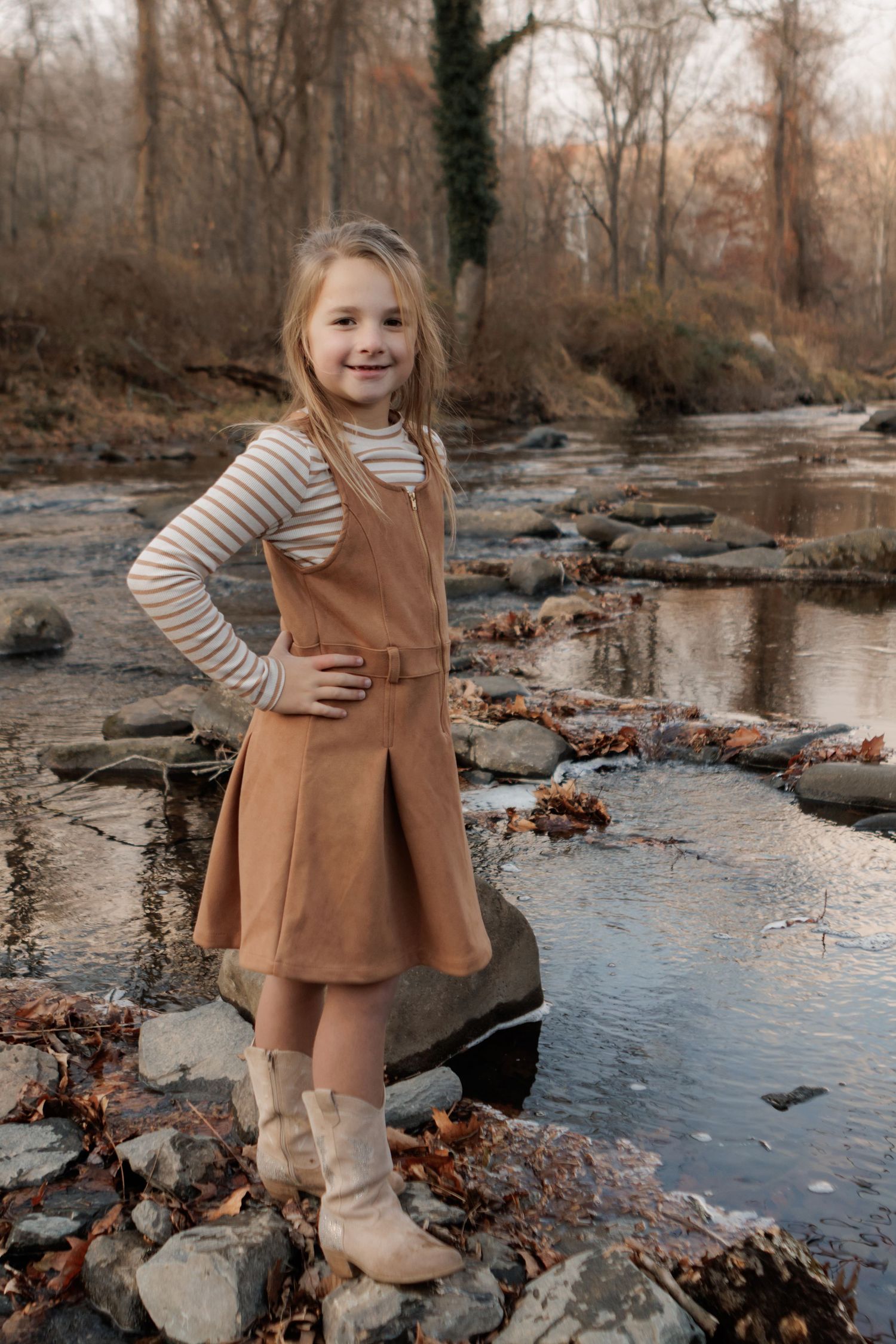Child in brown dress plays near stream with autumn leaves and rocks.
