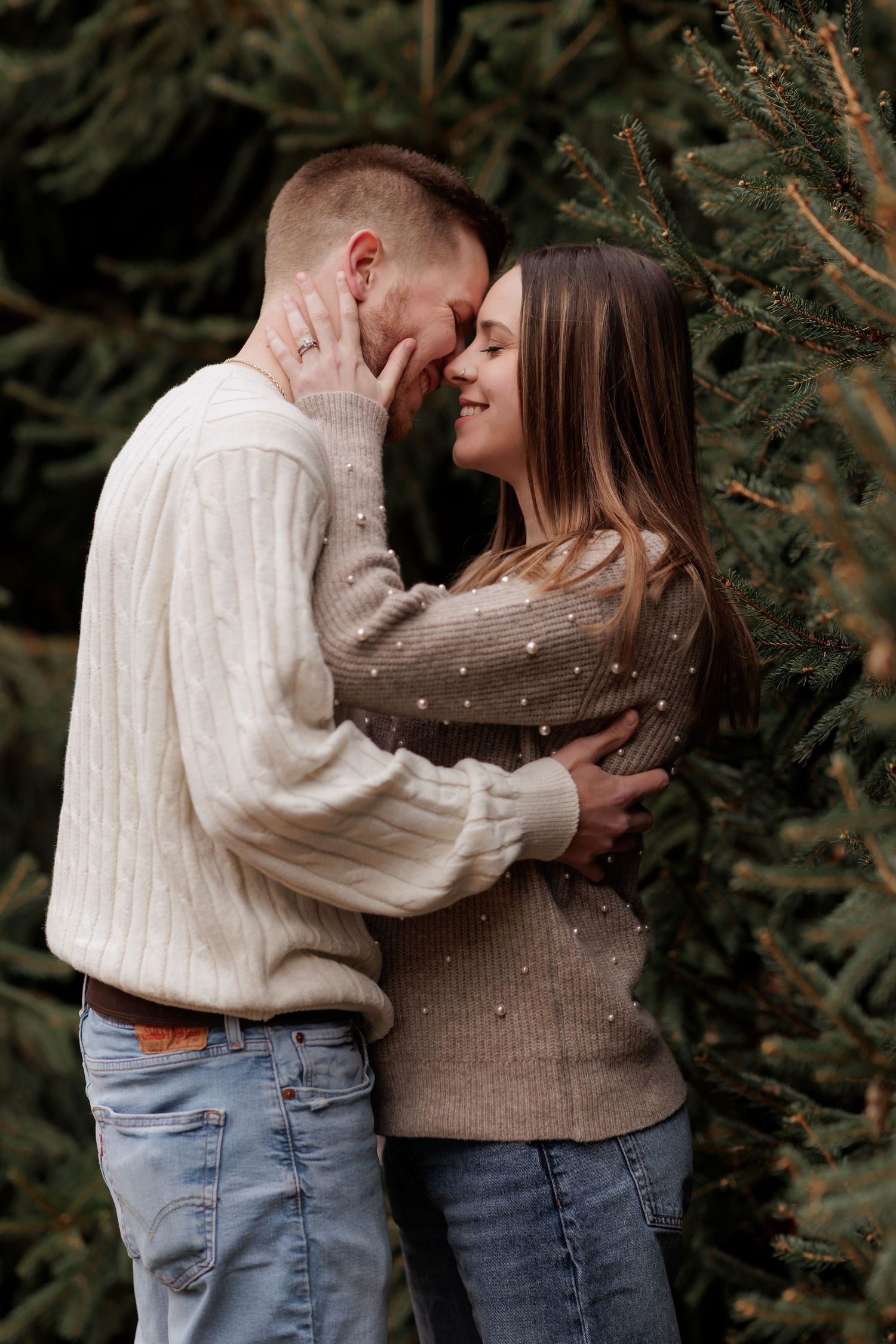 A couple in casual winter wear sharing an intimate moment near pine trees.
