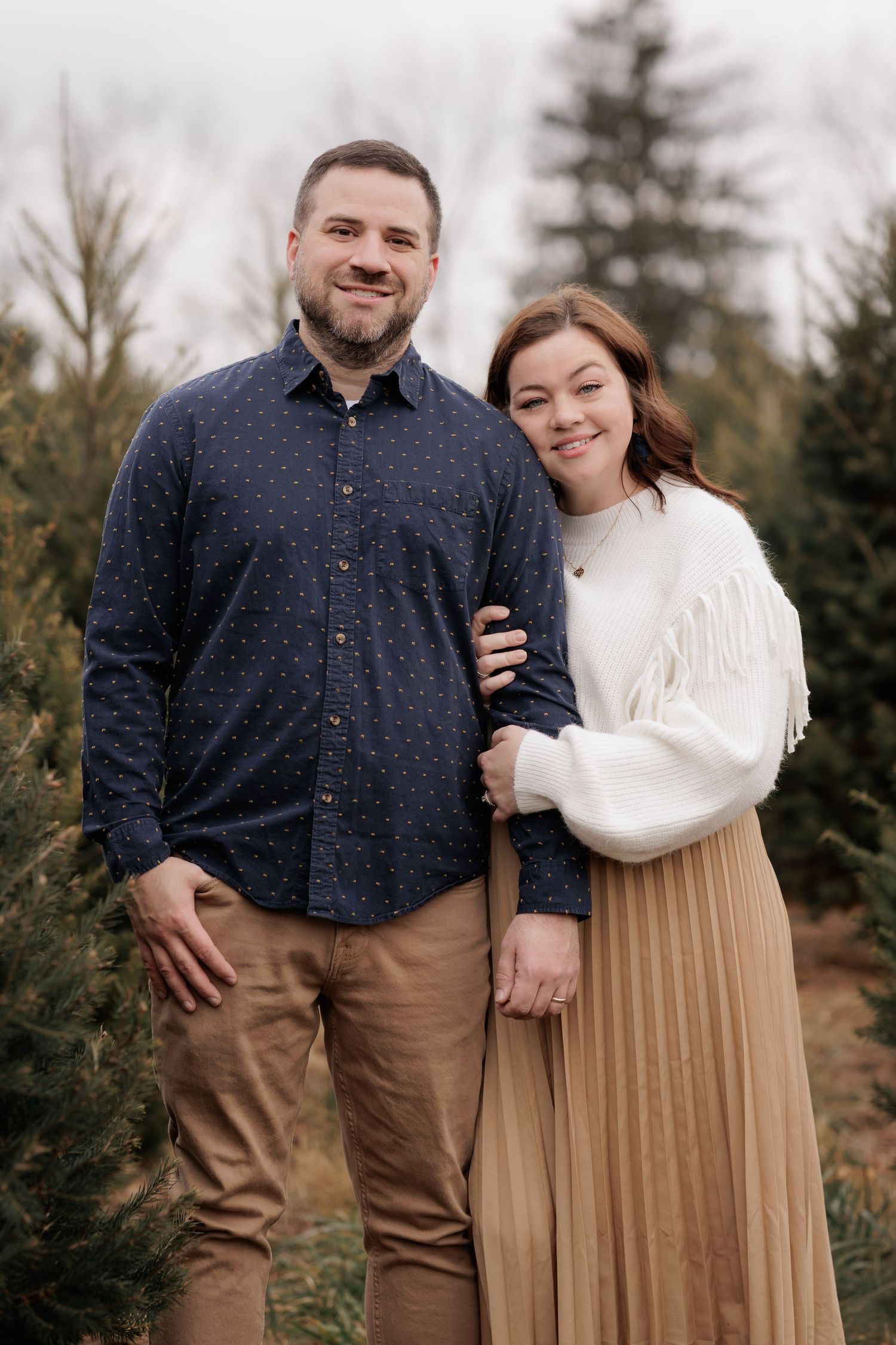 Couple poses together in casual attire against backdrop of evergreen trees.