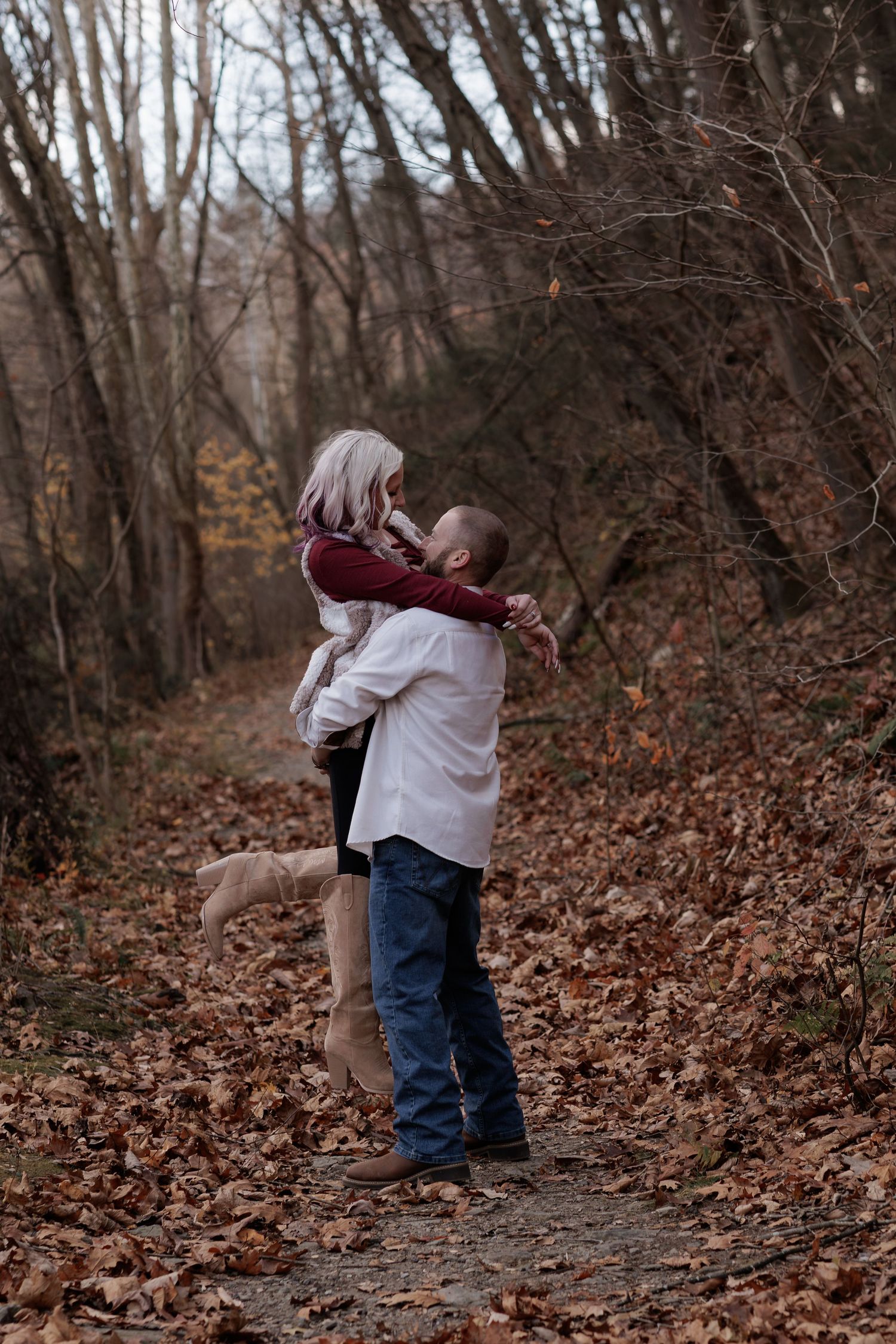 A couple embraces on a leaf-covered path in a wooded autumn setting.