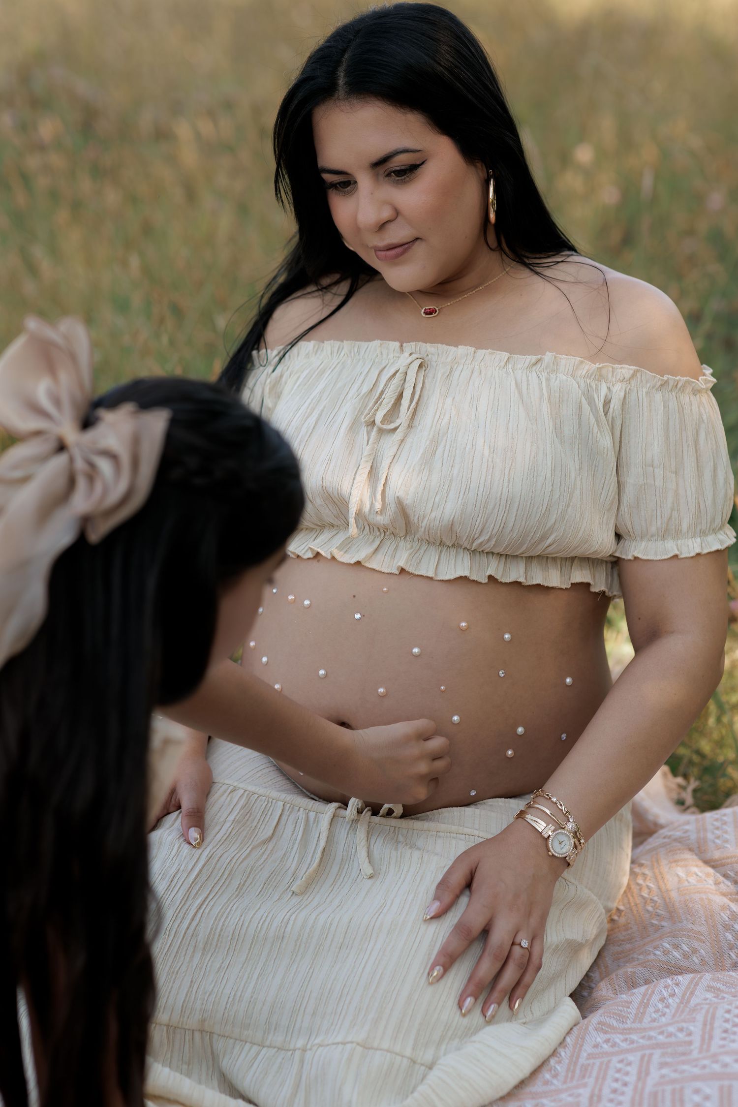 Pregnant woman in a cream off-shoulder top and skirt sits on a blanket in a grassy field, her belly decorated with pearls, beside a child.