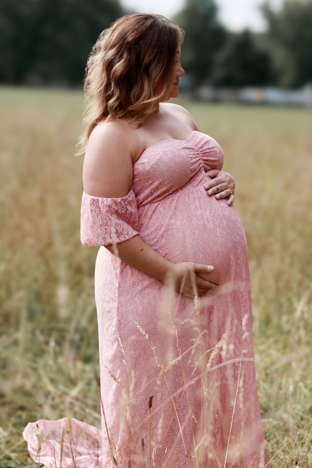 Pregnant woman in pink lace off-shoulder gown cradling her baby bump in a golden field.